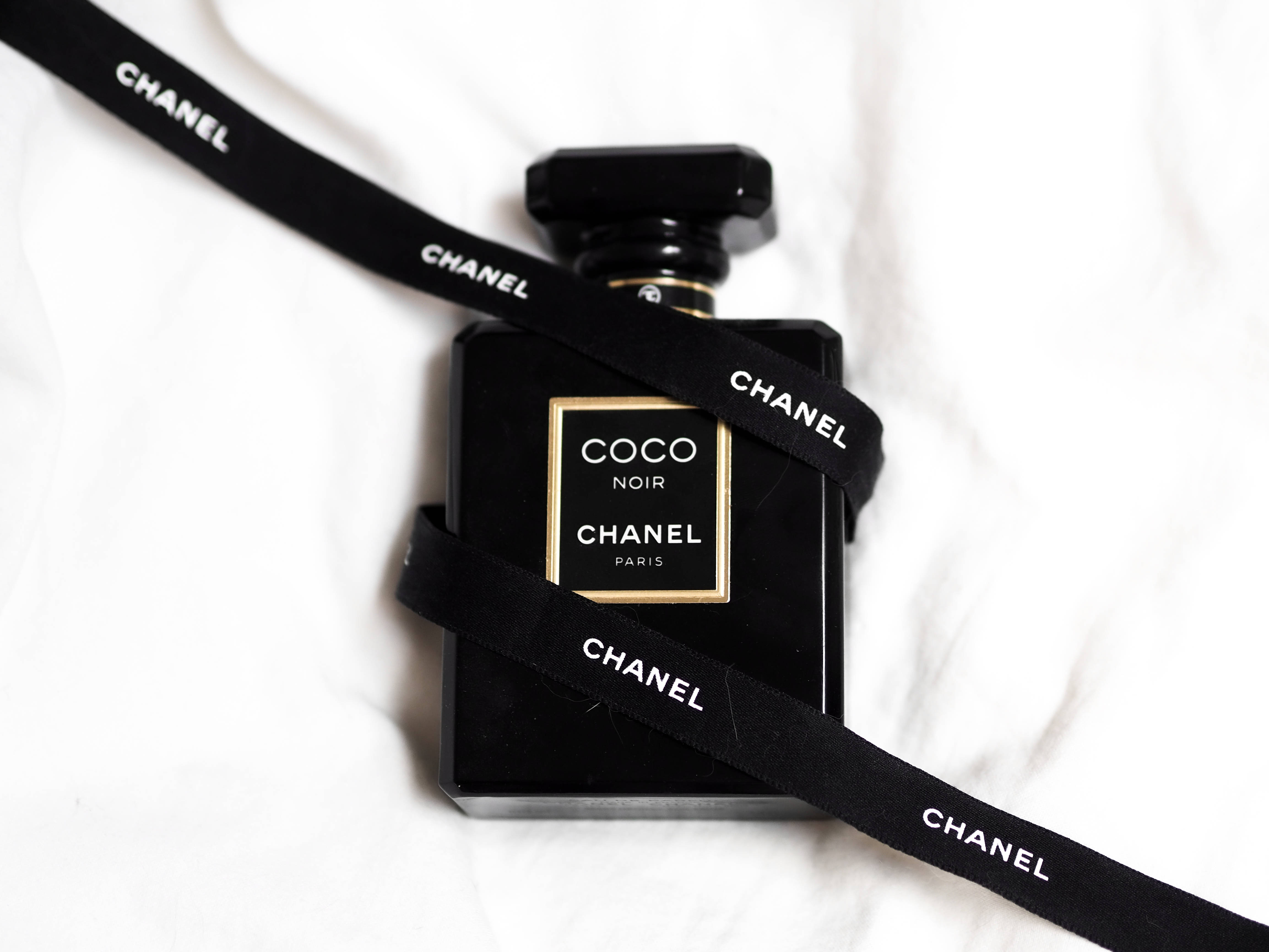 Chanel 桌布 壁紙 — 一個黑色 Chanel Coco Noir 香水瓶，包裹著黑色絲帶，帶有白色 Chanel 文字，放置在白色紋理布料上。