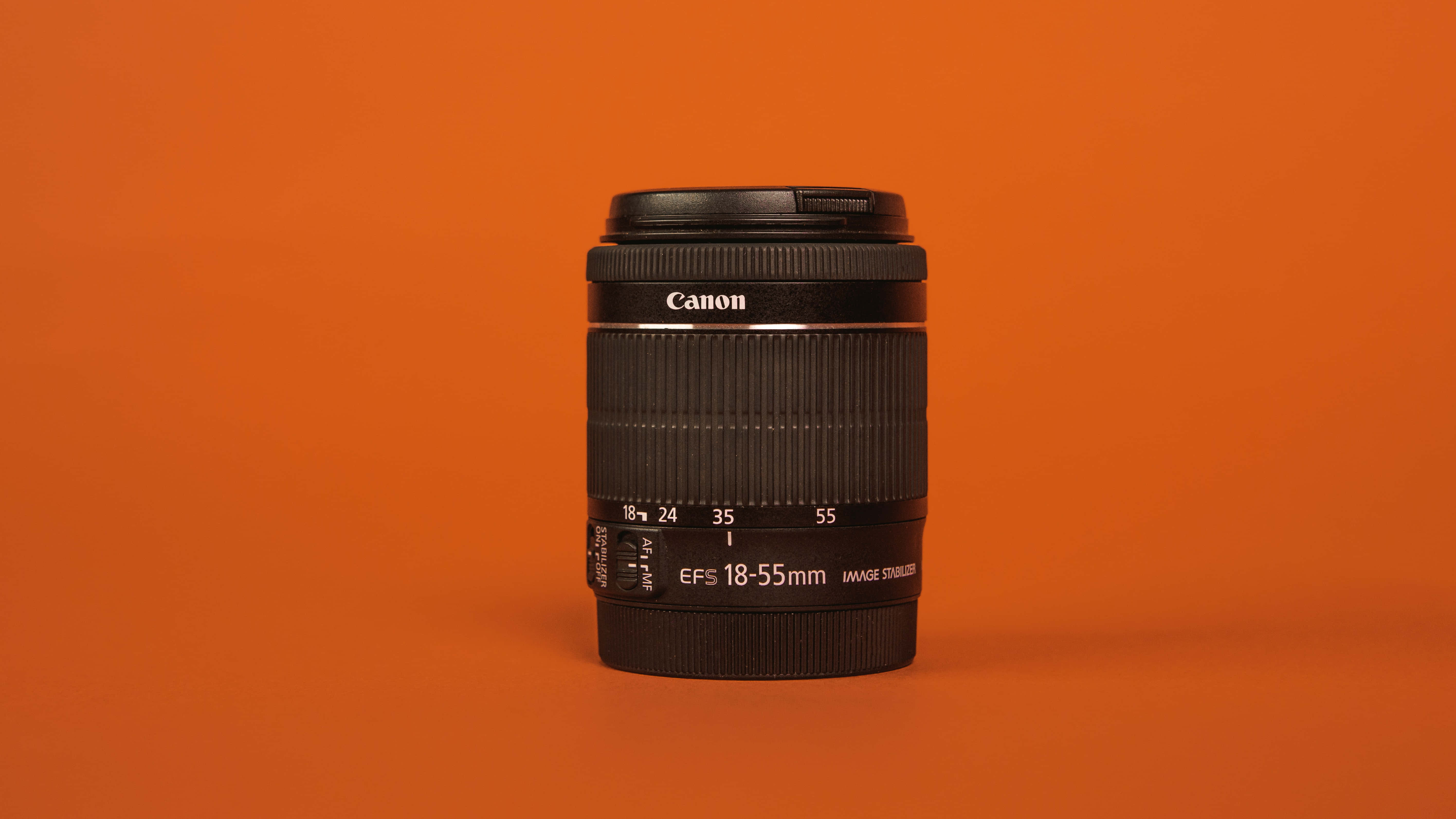 攝影 桌布 壁紙 — 一個黑色Canon EF-S 18-55mm相機鏡頭，居中放置在純色溫暖橙色背景上。