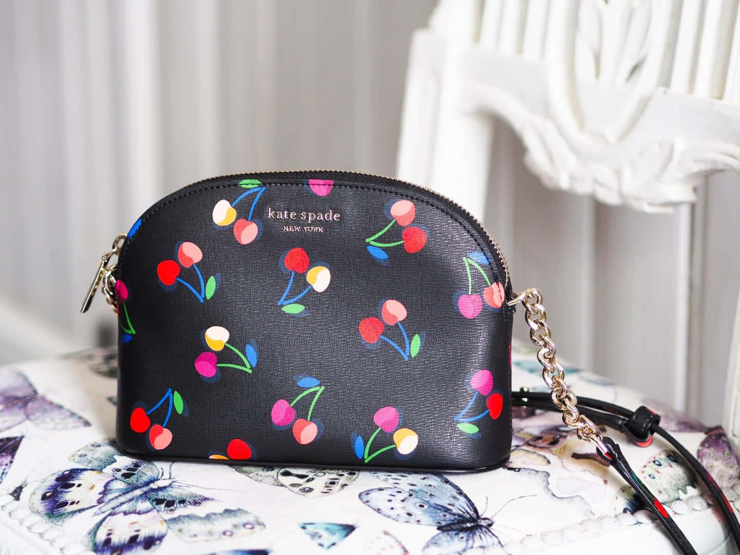 Kate Spade 桌布 壁紙 — 一個黑色Kate Spade斜背包，上面有色彩繽紛的櫻桃圖案，放在白色椅子附近的蝴蝶圖案布料上。