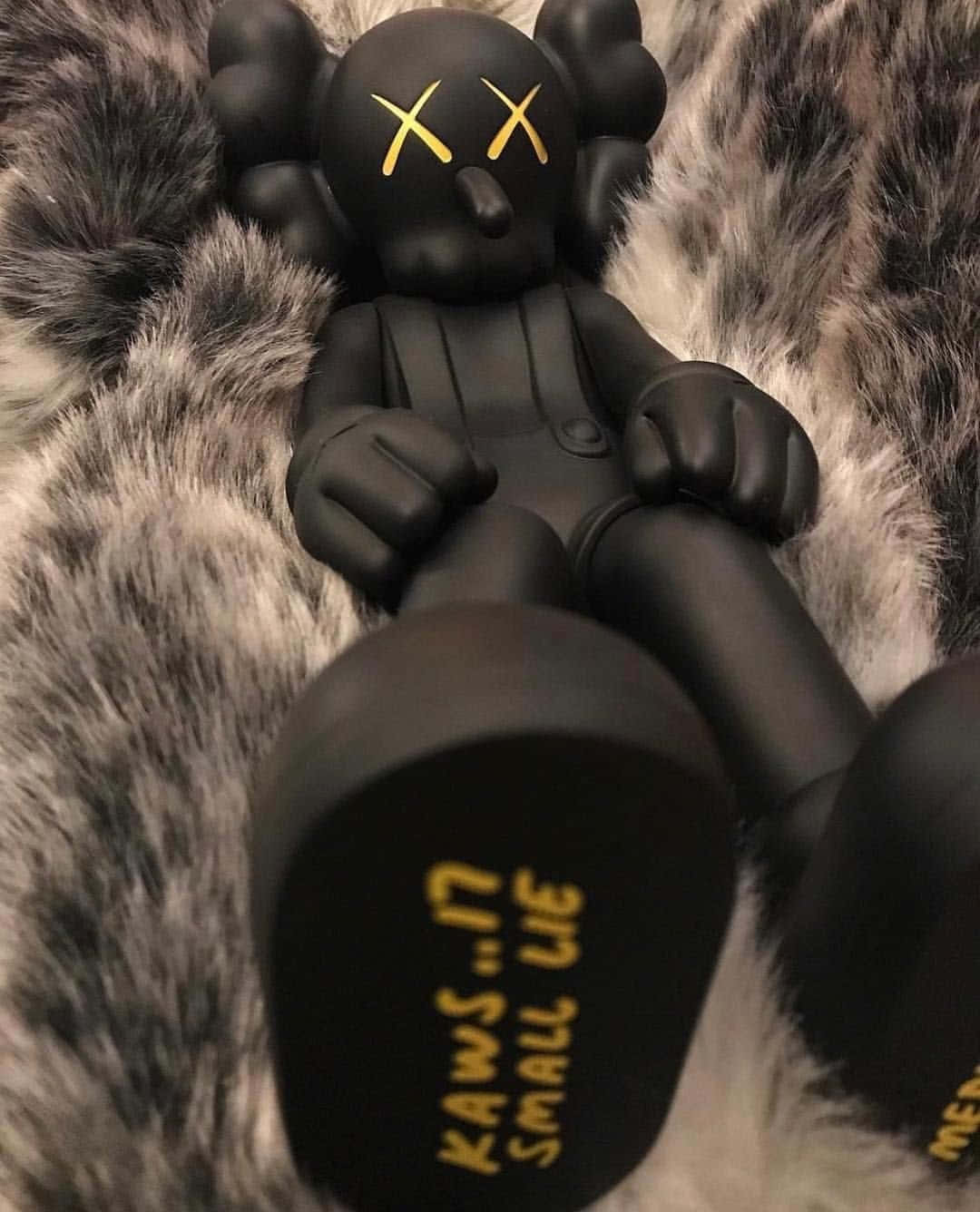 KAWS 桌布 壁紙 — 一個黑色KAWS Companion公仔，帶有黃色X眼睛，放在柔軟的灰白色毛絨表面上。