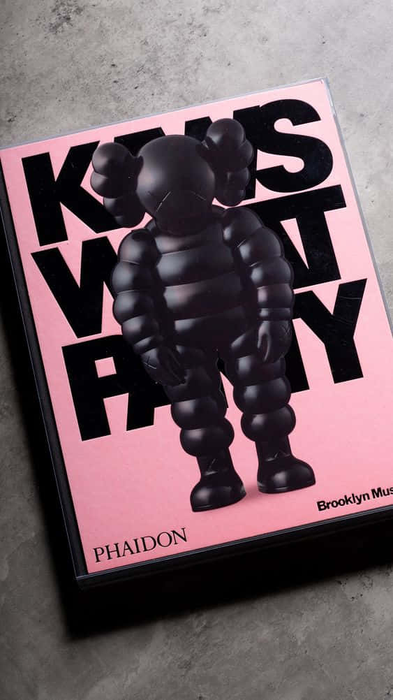 kaws pc 桌布 壁紙 — 一個黑色KAWS毛絨公仔站在粉紅色書封面上，書封面有粗體黑色文字，背景是灰色混凝土表面。