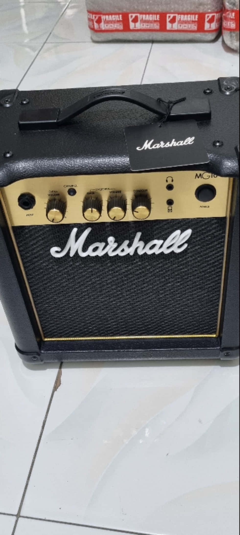 Marshall 桌布 壁紙 — 一個黑色Marshall MG10吉他擴音器，帶有金色飾邊，放在白色瓷磚地板上，旋鈕和標誌清晰可見。