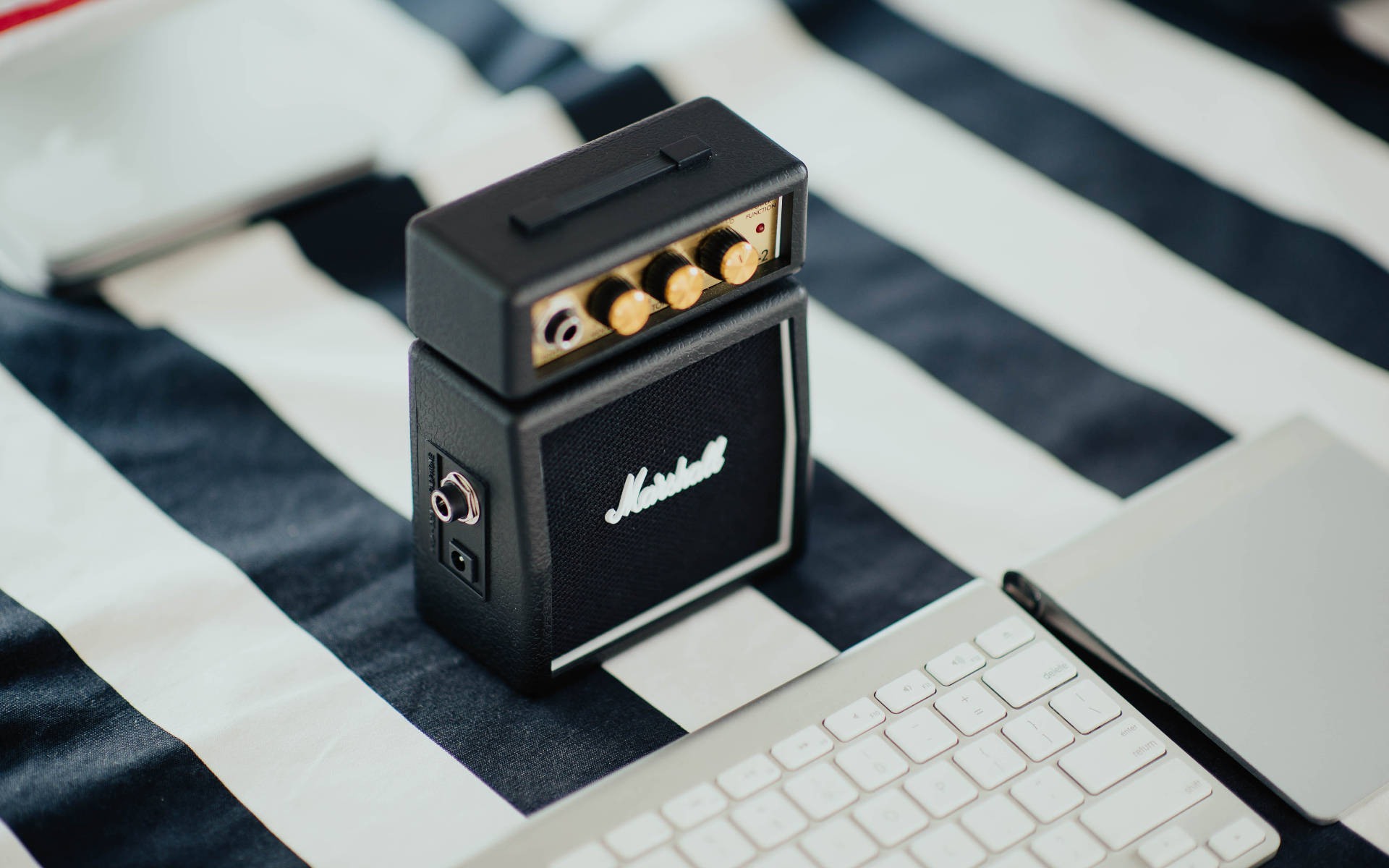 Marshall 桌布 壁紙 — 一個黑色Marshall迷你擴音器帶金色旋鈕，放在黑白條紋布料上，旁邊有一台白色筆記型電腦。