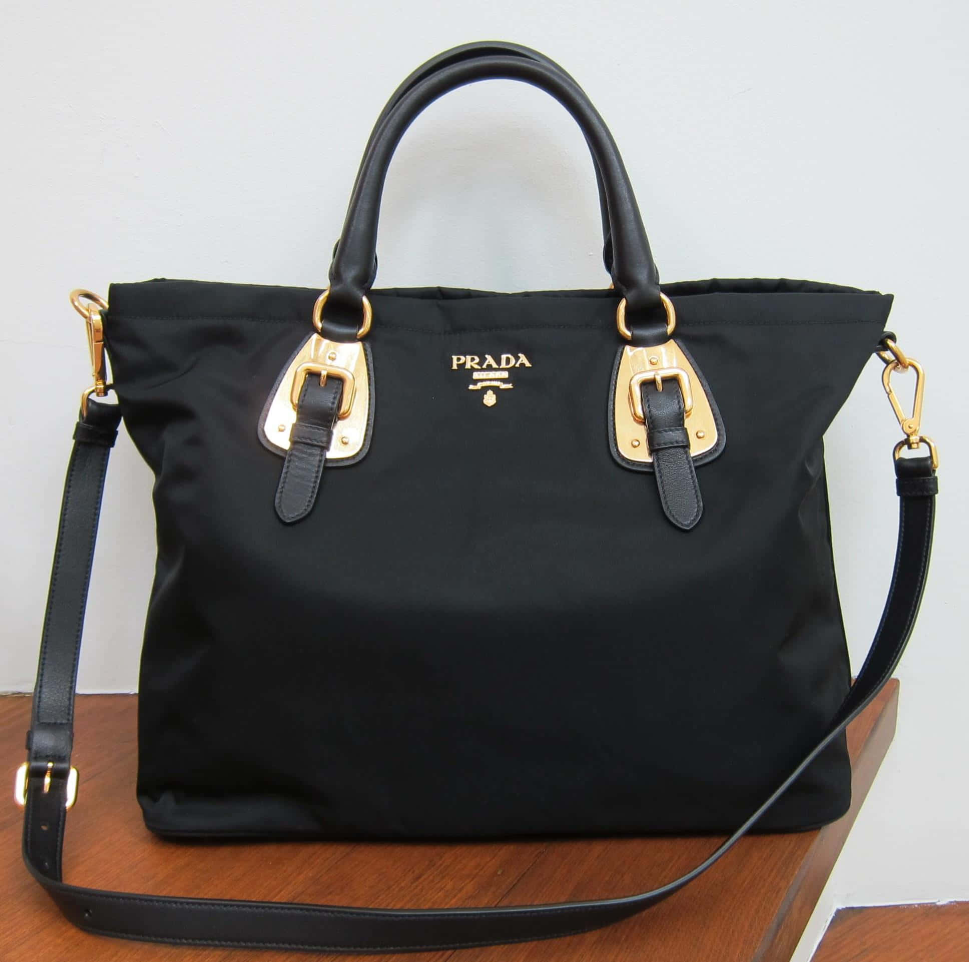 Prada 桌布 壁紙 — 一個黑色Prada托特包，配有金色金屬配件，放在木質表面上，背景是純白牆壁。