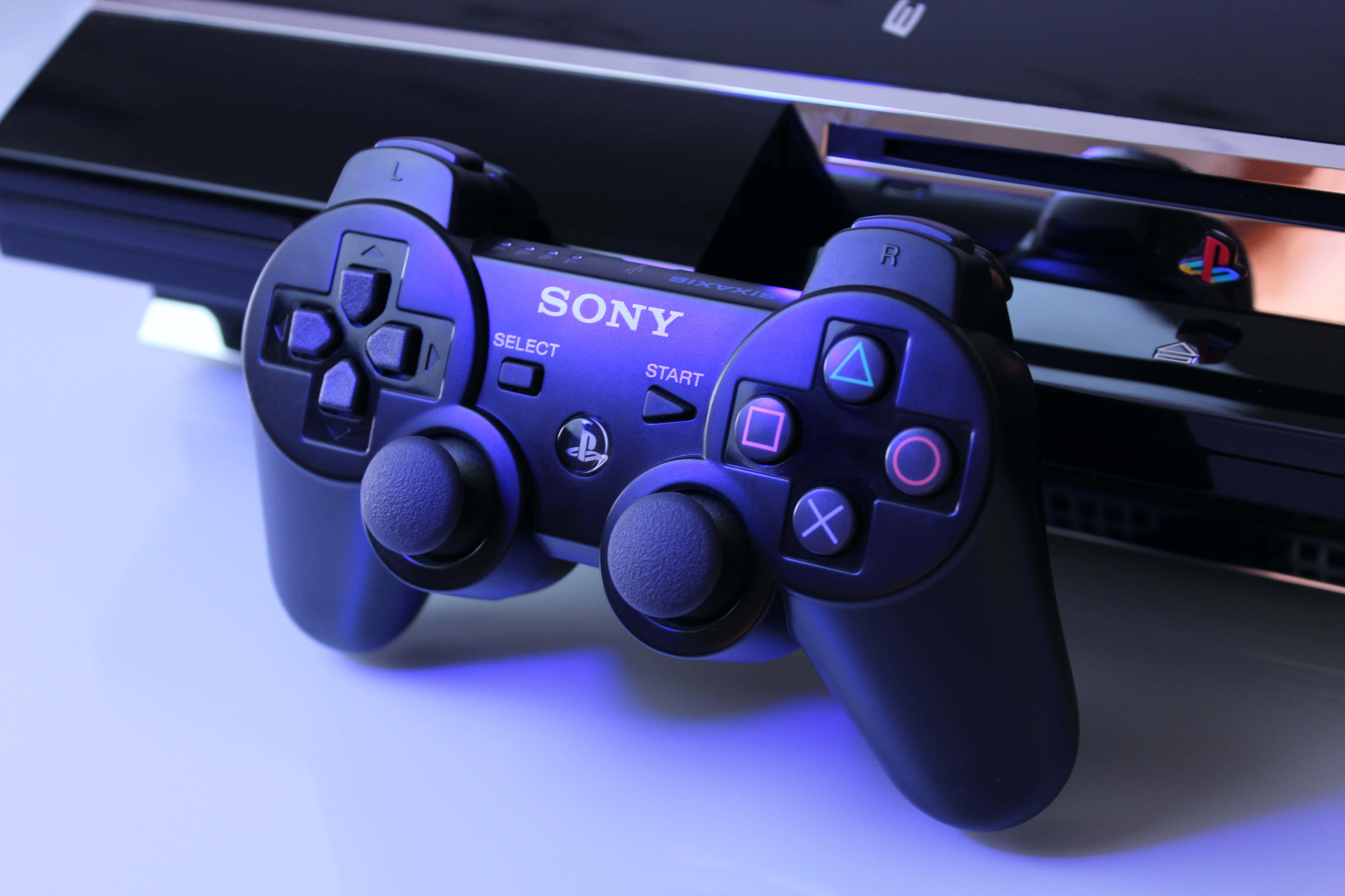 Gaming Consoles 桌布 壁紙 — 一個黑色Sony PlayStation 4控制器在藍光下發光，旁邊是一個白色表面上的主機。