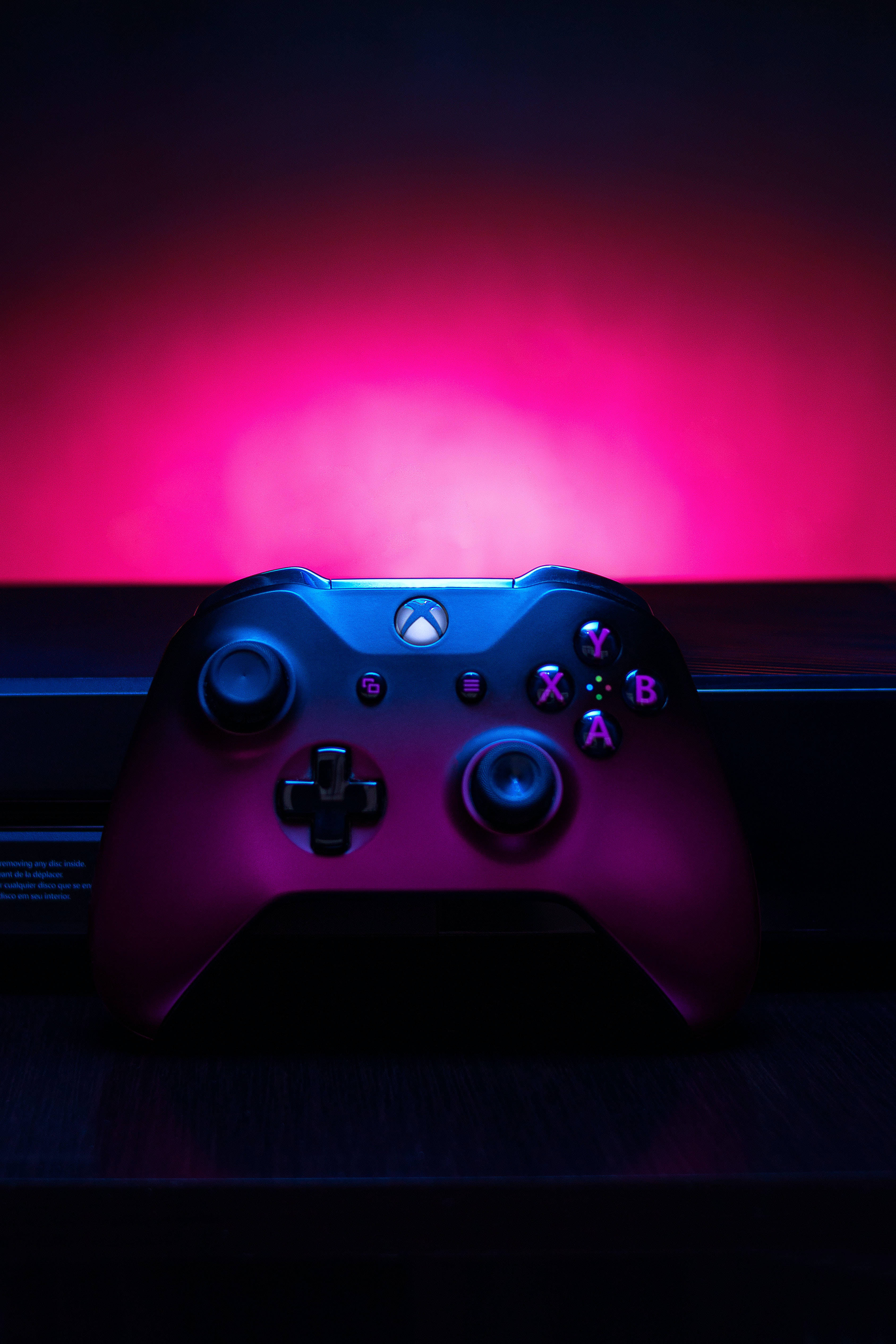 Dark Purple iPhone 桌布 壁紙 — 一個黑色Xbox手把在深色背景上沐浴在充滿活力的粉紅和藍色霓虹燈光中。