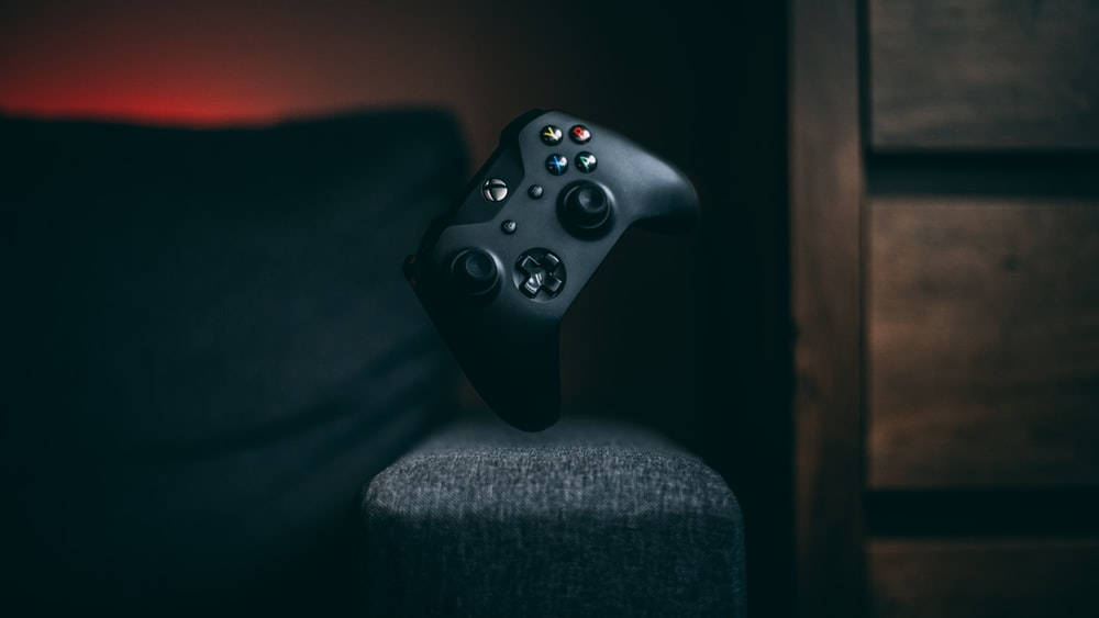 遊戲 桌布 壁紙 — 一個黑色Xbox手把放在灰色織物扶手上，背景為深色木材，帶有溫暖的環境燈光。