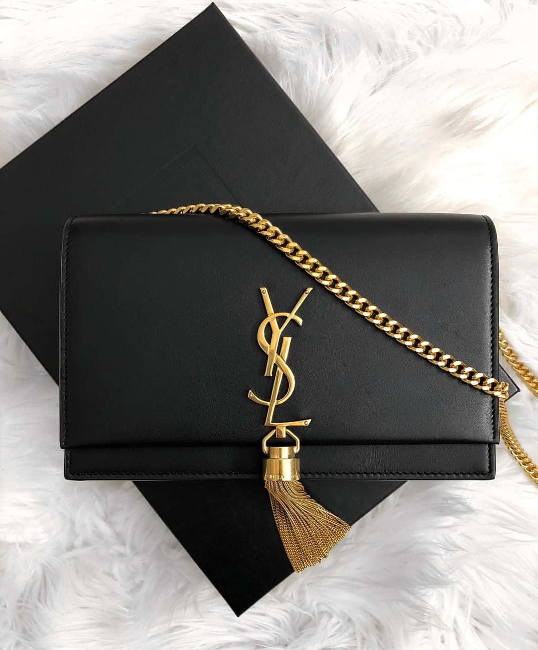 YSL 桌布 壁紙 — 一個黑色YSL手袋，配有金色鏈條和流蘇，優雅地放置在毛絨白色表面上，旁邊是它的黑色盒子。