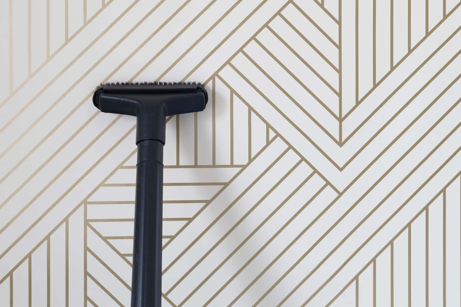 Cleaning 桌布 壁紙 — 一個黑色吸塵器頭靠在一面帶有金色幾何V形圖案的白色牆上。