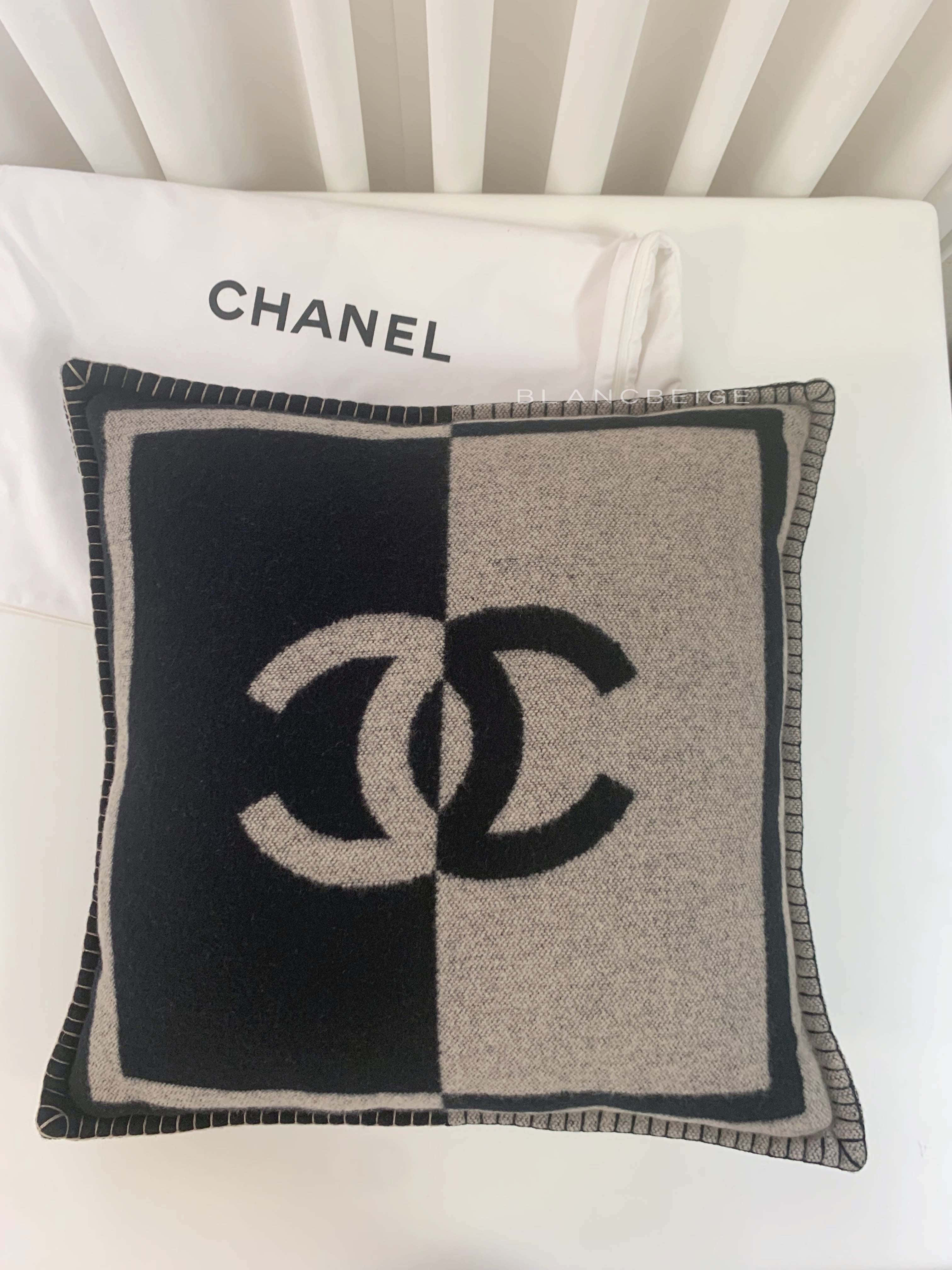 香奈兒標誌 桌布 壁紙 — 一個黑色和米色的 Chanel 標誌枕頭放在一個帶有 'CHANEL' 文字的白色枕頭上，背景是白色板條床頭板。