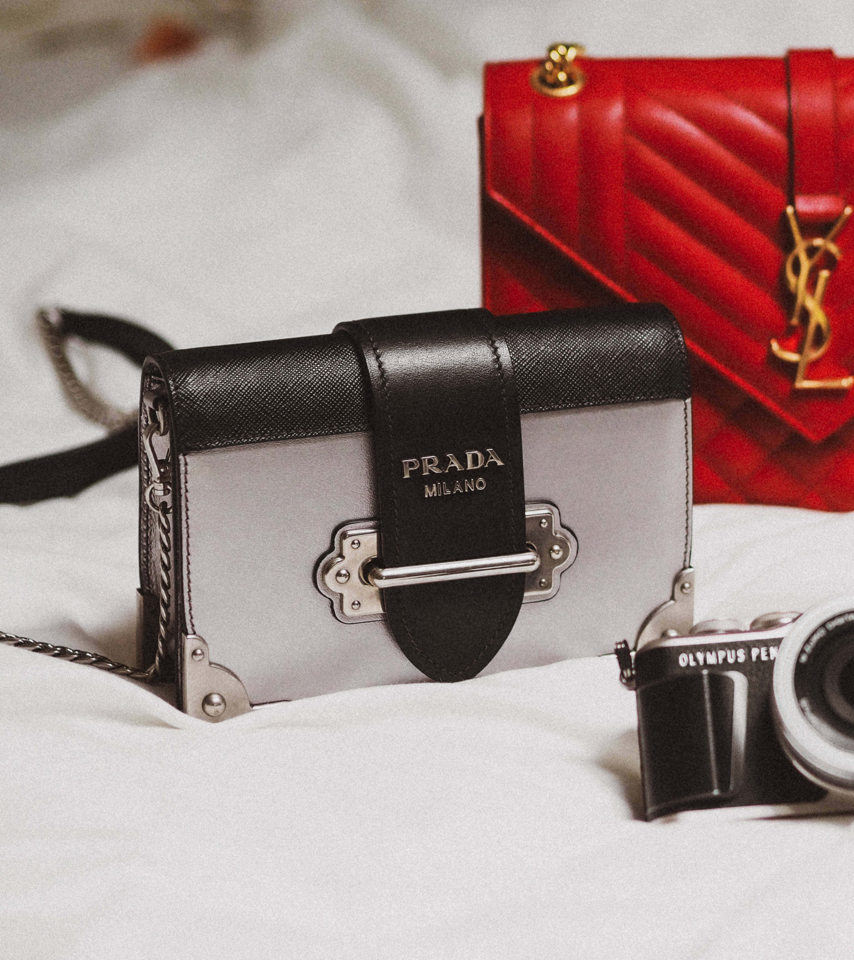 Gucci 桌布 壁紙 — 一個黑色和銀色的Prada手提包和紅色絎縫YSL手提包放在白色布料表面上的黑色Olympus相機旁邊。
