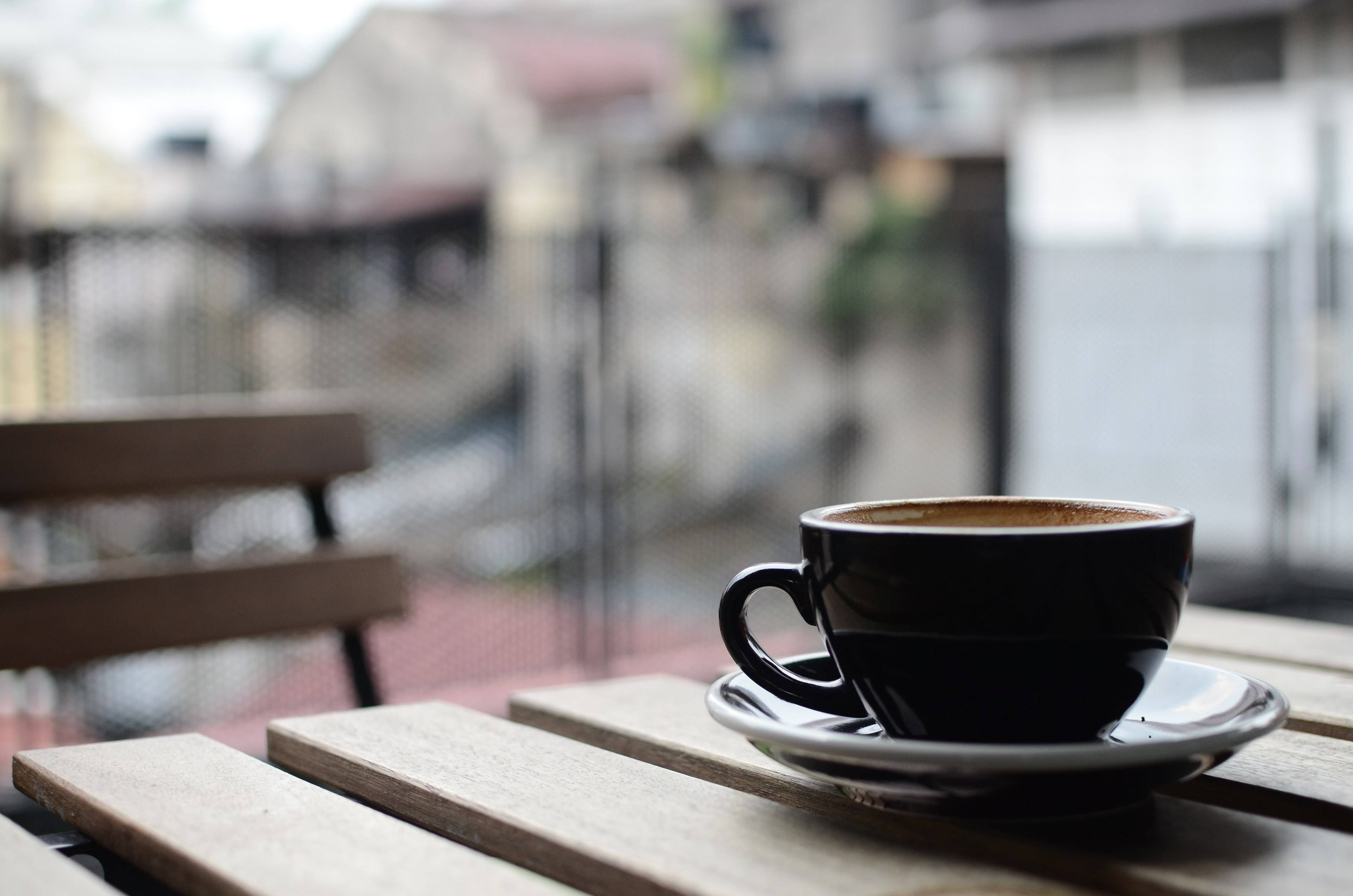 Coffee Aesthetic 桌布 壁紙 — 一個黑色咖啡杯放在戶外木桌上，背景是模糊的城市景觀。