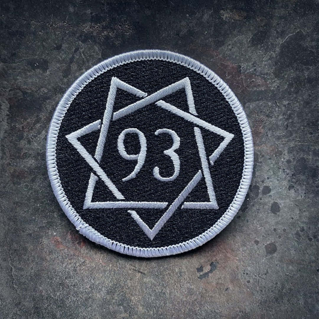 Thelema 桌布 壁紙 — 一個黑色圓形刺繡補丁，帶有白色縫線，在紋理灰色表面上顯示幾何星形圖案內的數字93。