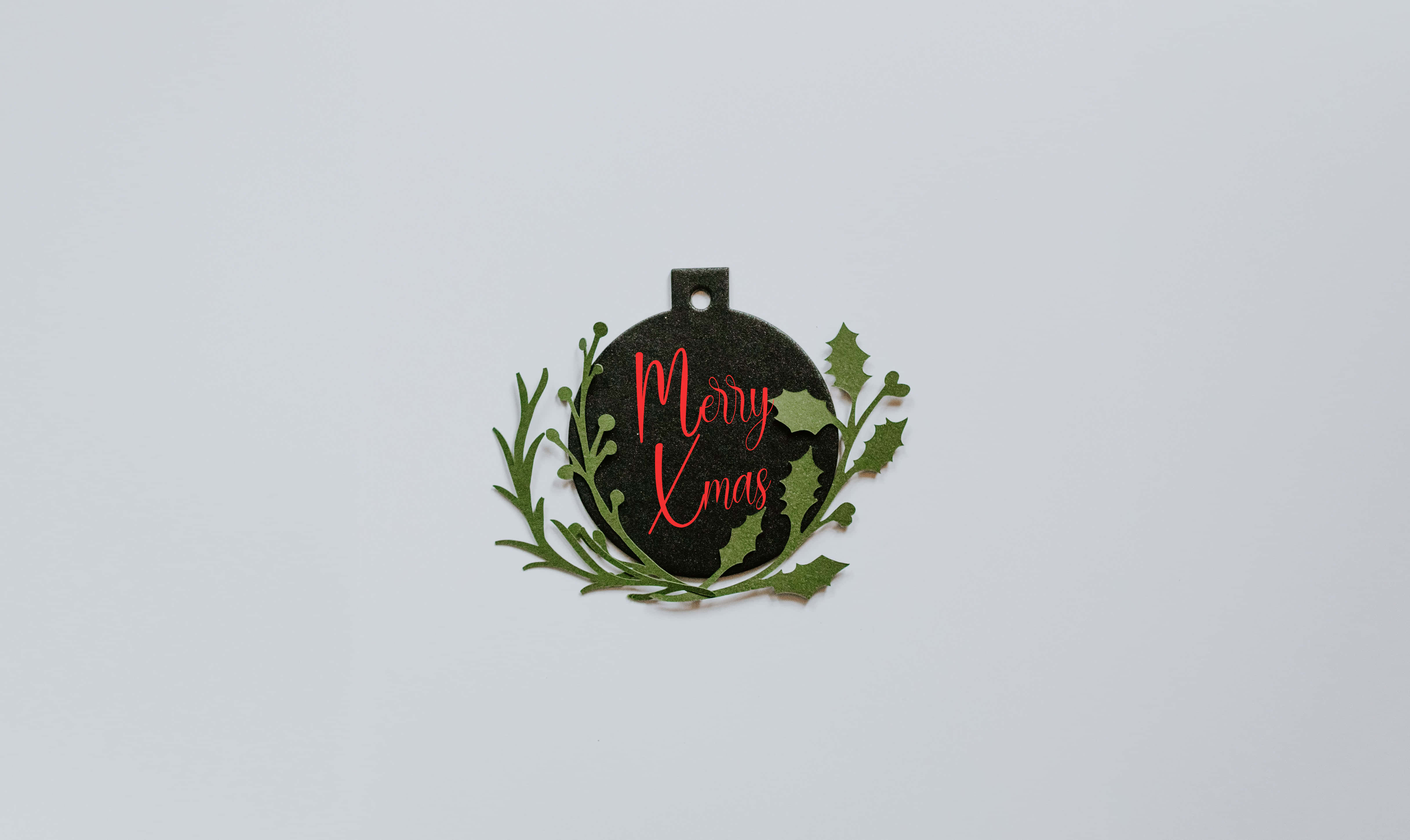Christmas Ball 桌布 壁紙 — 一個黑色圓形聖誕裝飾，上面有紅色草書 Merry Xmas 文字和綠色冬青葉，背景為純白色。