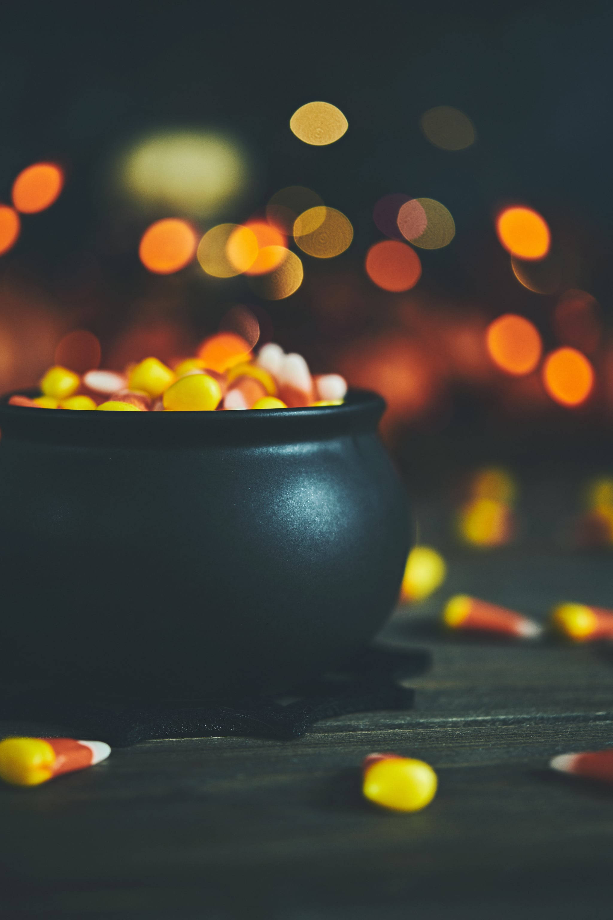 Cute Halloween Phone 桌布 壁紙 — 一個黑色大鍋裝滿了糖果玉米，放在深色木質表面上，背景有溫暖的散景燈光。