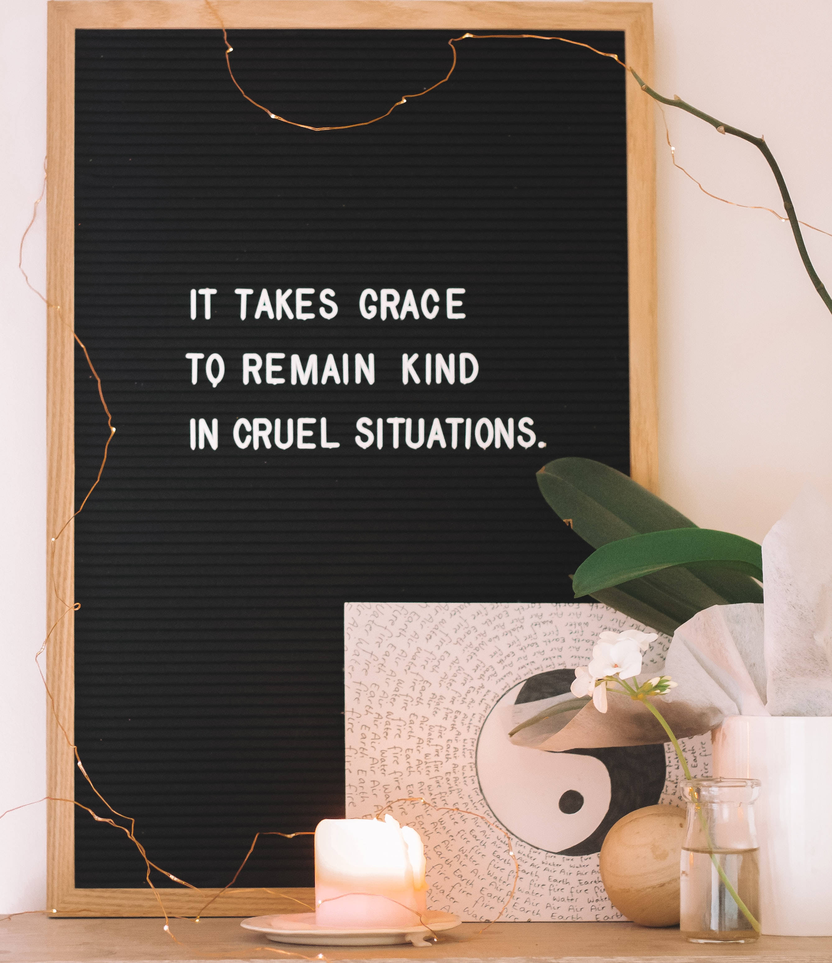 激勵 桌布 壁紙 — 一個黑色字母板，上面有白色文字寫著「IT TAKES GRACE TO REMAIN KIND IN CRUEL SITUATIONS」，周圍環繞著點燃的蠟燭、綠色植物和木架上的燈串。