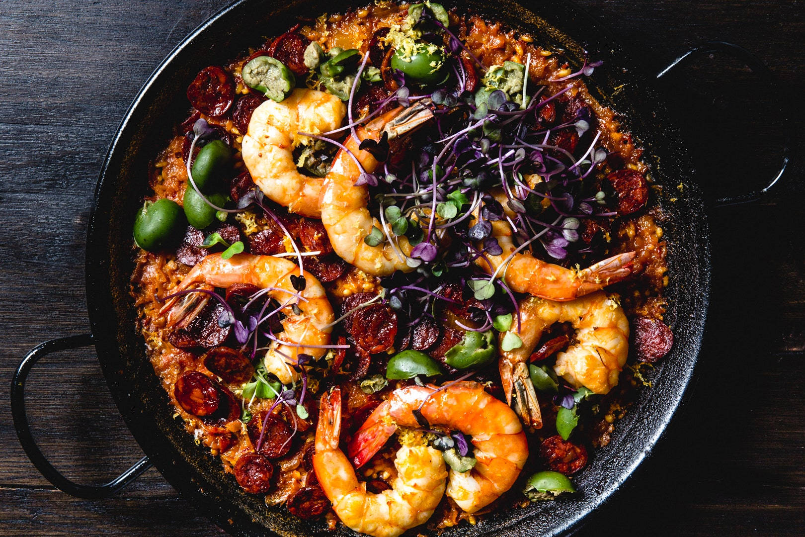 prawn 桌布 壁紙 — 一個黑色平底鍋中的色彩繽紛西班牙海鮮飯，上面鋪有蝦、西班牙辣香腸、青椒和紫色微型蔬菜，放在深色木質表面上。