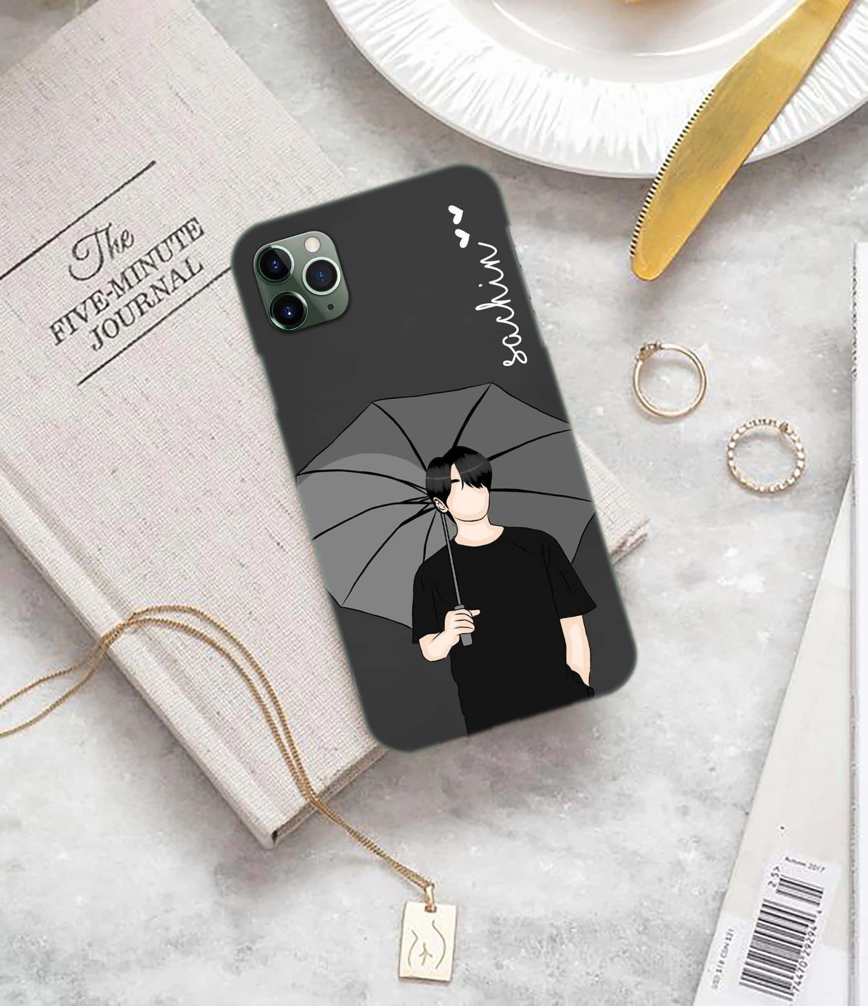 Phone Case 桌布 壁紙 — 一個黑色手機殼，上面有簡約動漫風格男孩撐著雨傘，旁邊放著日記本和金飾，置於大理石表面上。