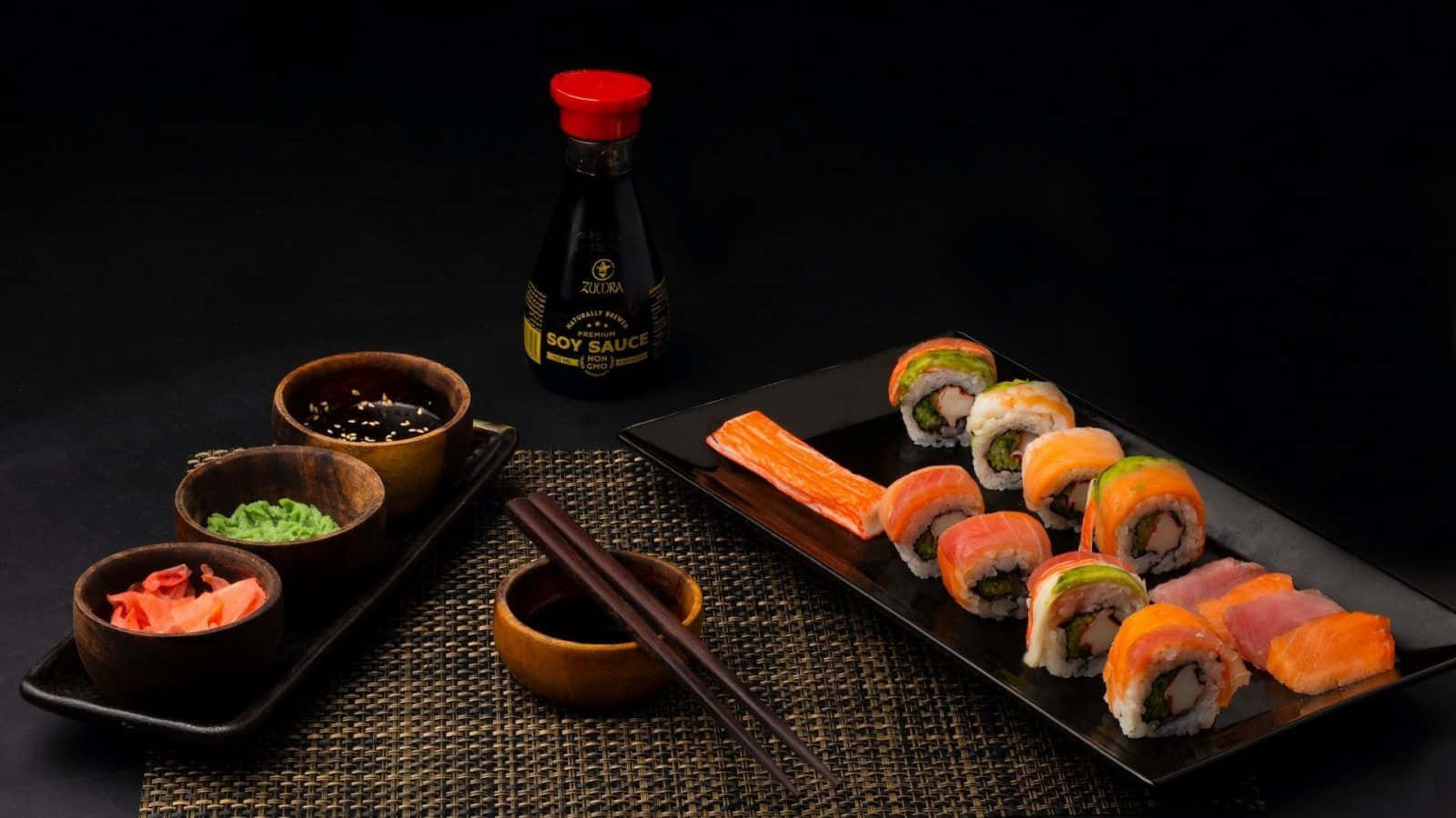 Sushi 桌布 壁紙 — 一個黑色托盤上放著色彩繽紛的壽司卷和生魚片，旁邊有醬油、生薑、芥末和筷子，置於深色編織墊上。