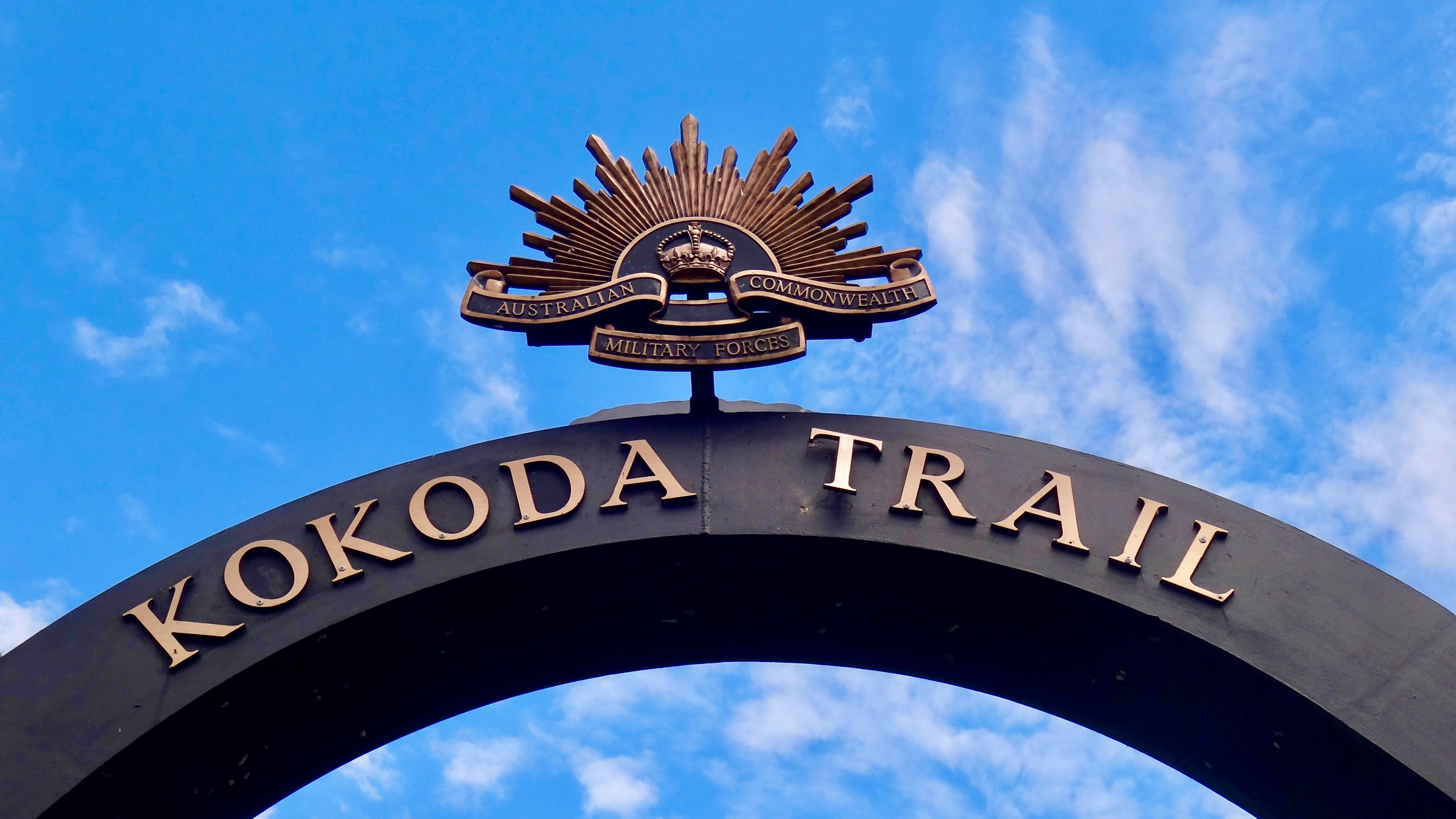 New 桌布 壁紙 — 一個黑色拱門，上面有金色字體寫著Kokoda Trail，頂部有澳洲聯邦軍隊徽章，背景是明亮藍天和縷縷白雲。