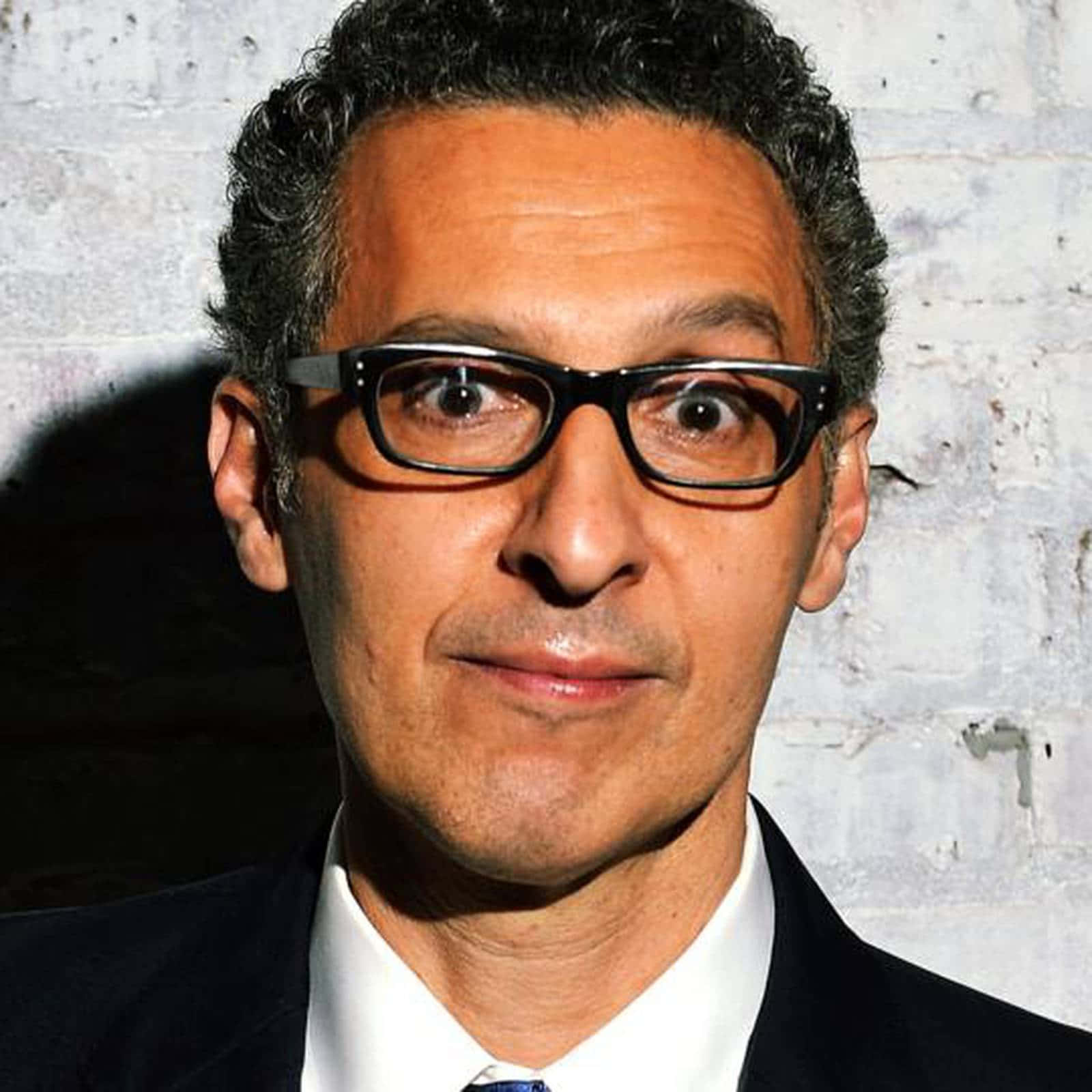 John Turturro 桌布 壁紙 — 一個黑色捲髮、戴黑框眼鏡的男人，在風化的白色磚牆前專注地凝視鏡頭。