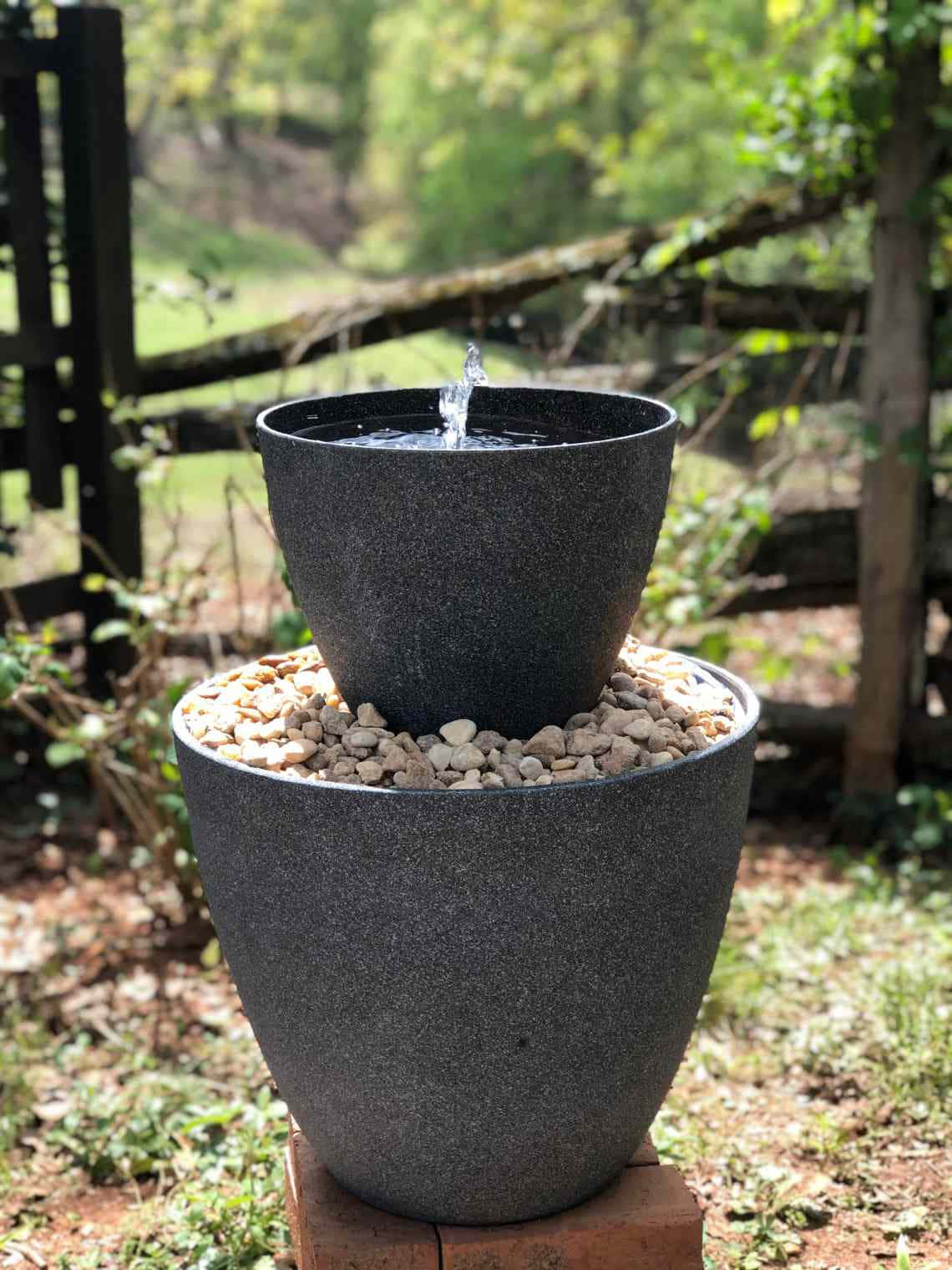 Outdoor 桌布 壁紙 — 一個黑色斑點兩層戶外噴泉，水從頂部到底部層層流下，周圍環繞著鵝卵石和綠色植物。