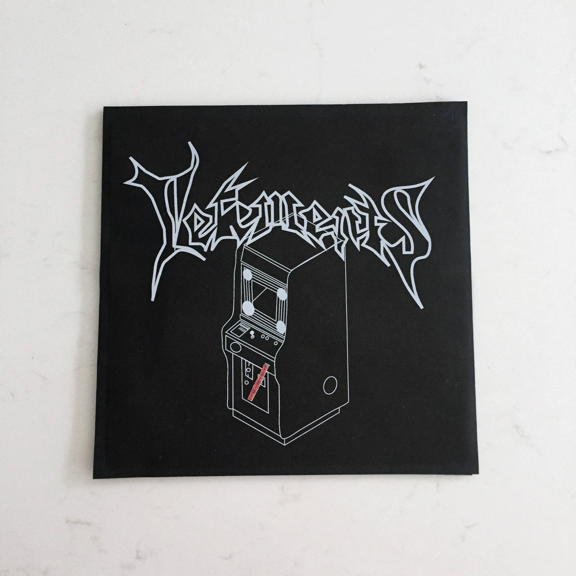 Vetements 桌布 壁紙 — 一個黑色方形貼紙，上面有白色塗鴉風格文字和簡約街機輪廓，放在大理石表面上。