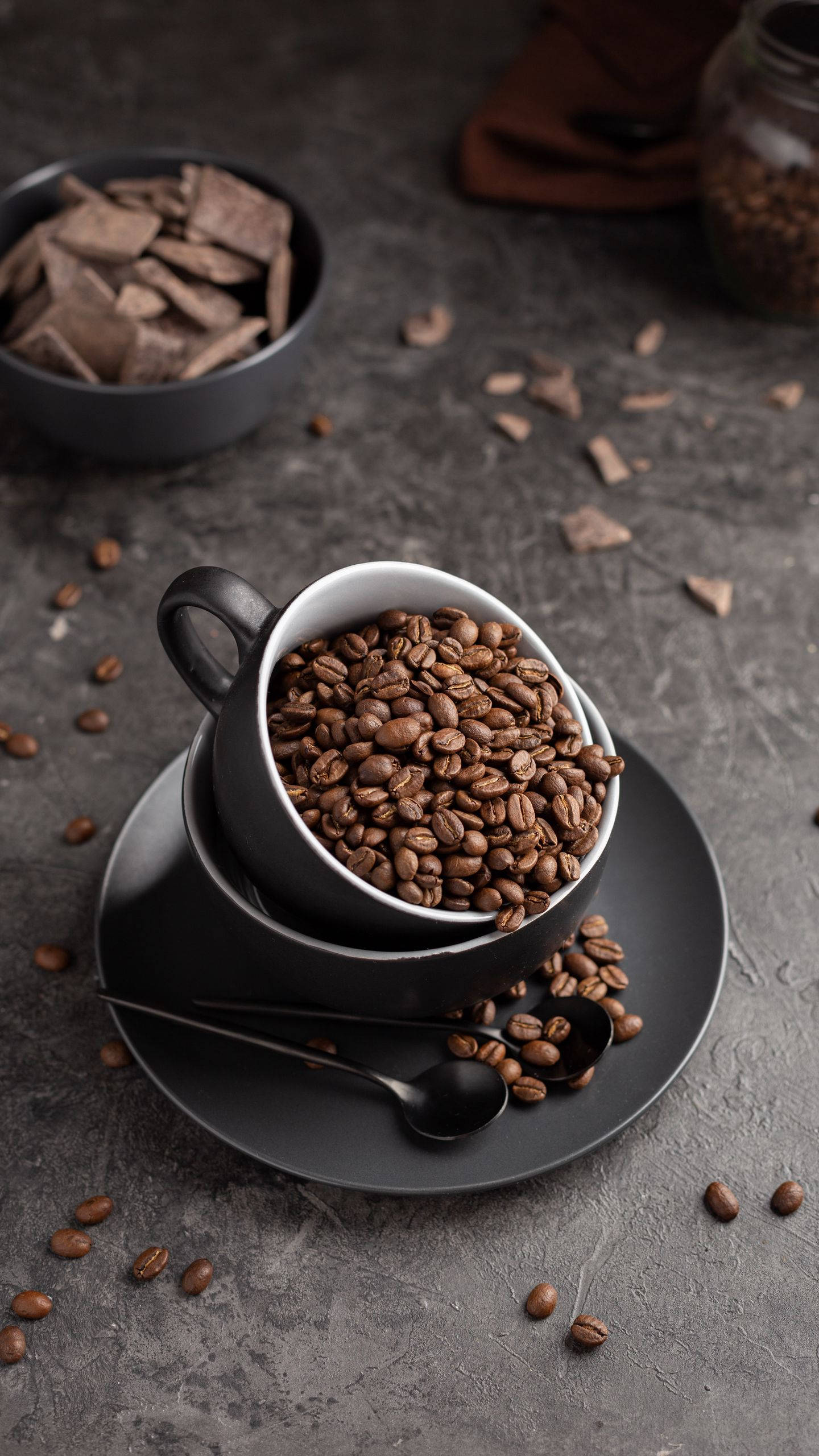beans 桌布 壁紙 — 一個黑色杯子溢出烘焙咖啡豆，放在碟子上，周圍散落著豆子和巧克力塊，置於紋理灰色表面上。