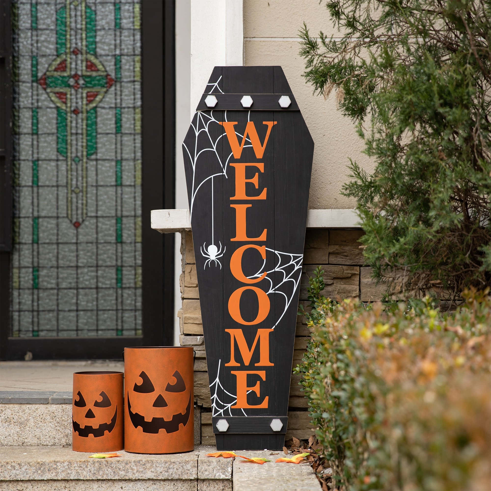 Halloween Coffins 桌布 壁紙 — 一個黑色棺材形狀的歡迎標誌，帶有橙色字母和白色蜘蛛網，與南瓜燈籠桶一起立在石頭門廊上。