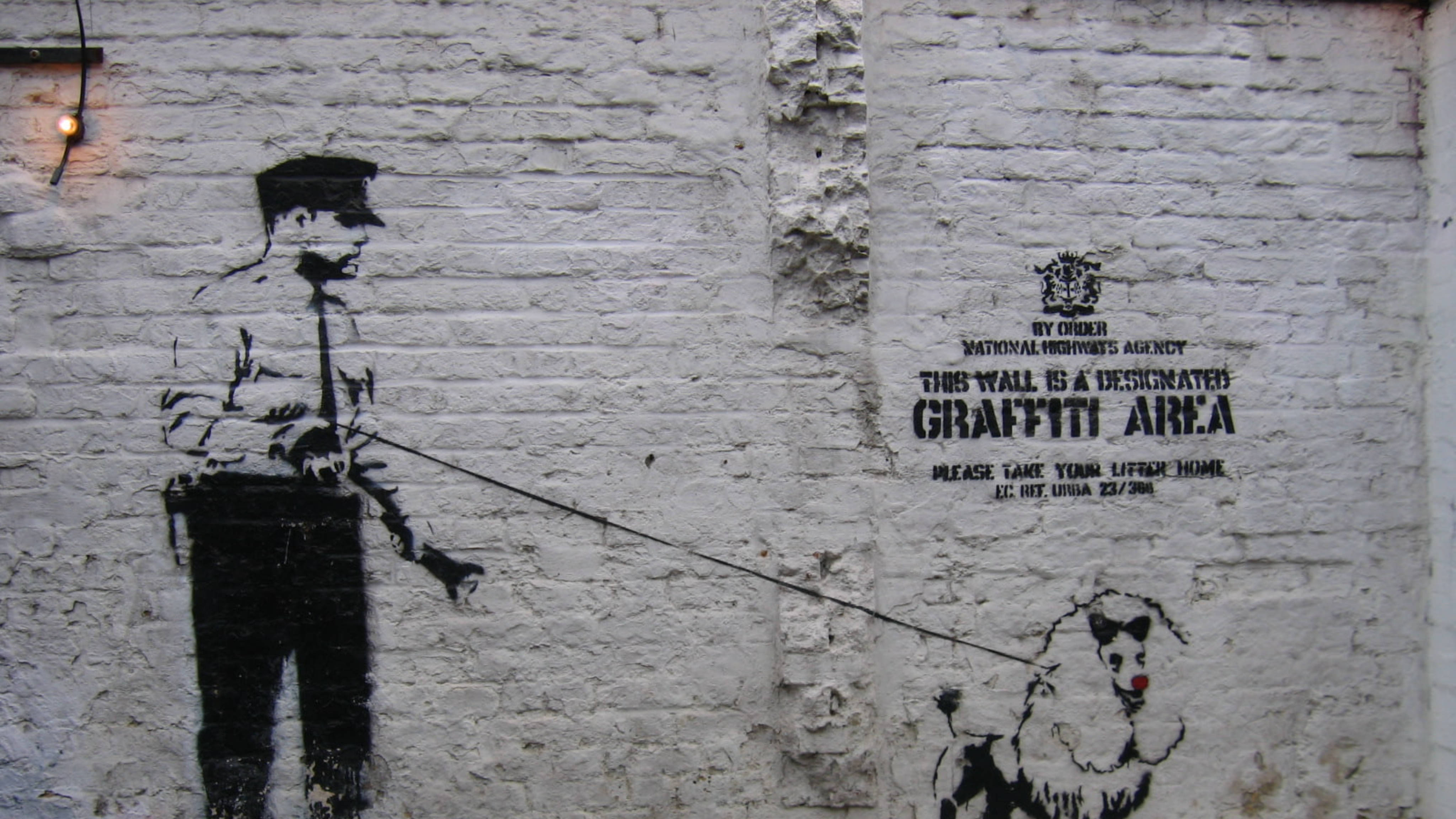 Banksy 桌布 壁紙 — 一個黑色模板塗鴉，描繪一名警察在白色磚牆上，牽著一條狗，狗身上標有「此牆為指定塗鴉區」。
