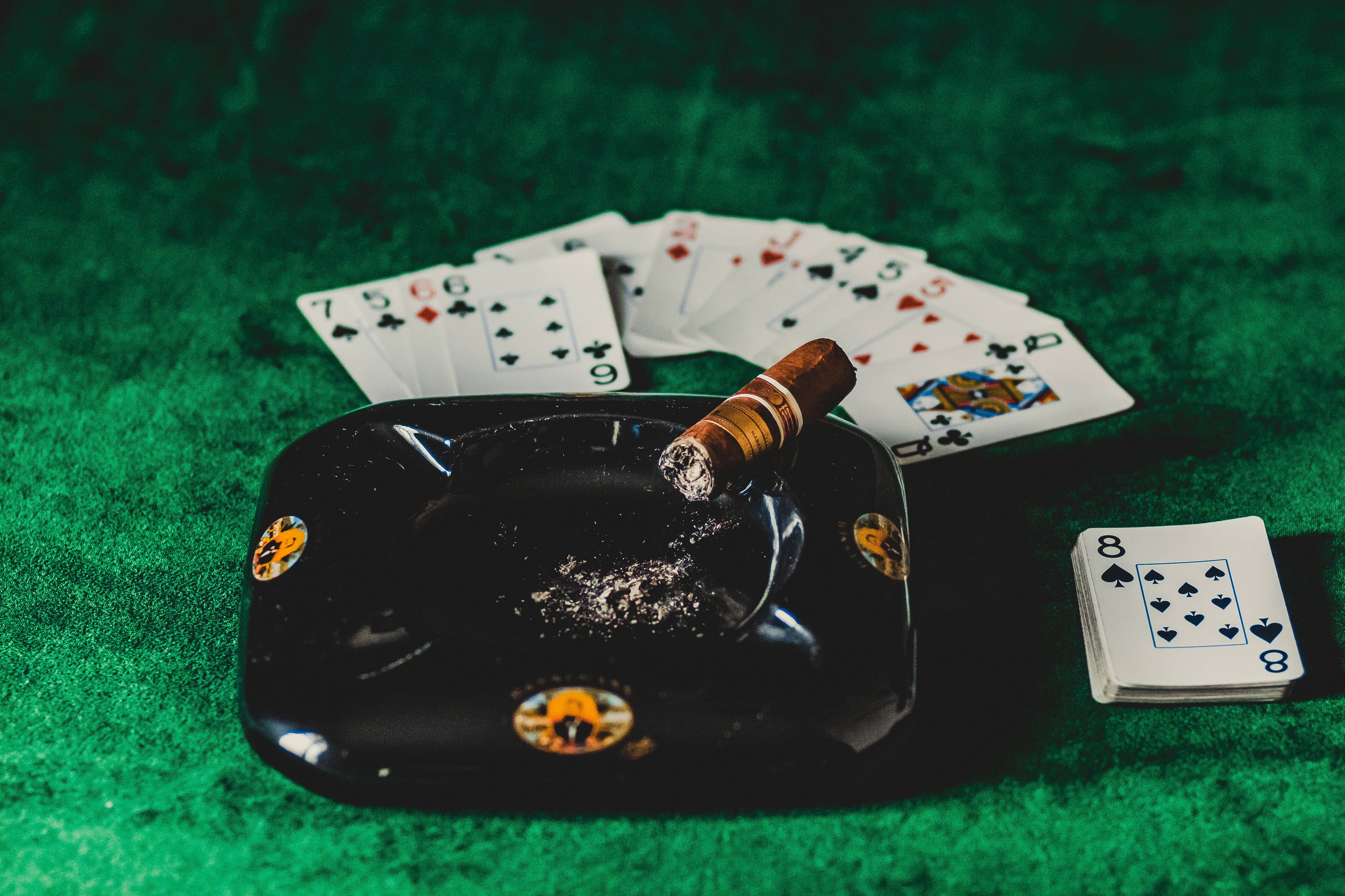 Casino 桌布 壁紙 — 一個黑色煙灰缸上放著一支點燃的雪茄和散落的籌碼，擱在綠色絨布桌上，旁邊是展開成扇形的撲克牌。