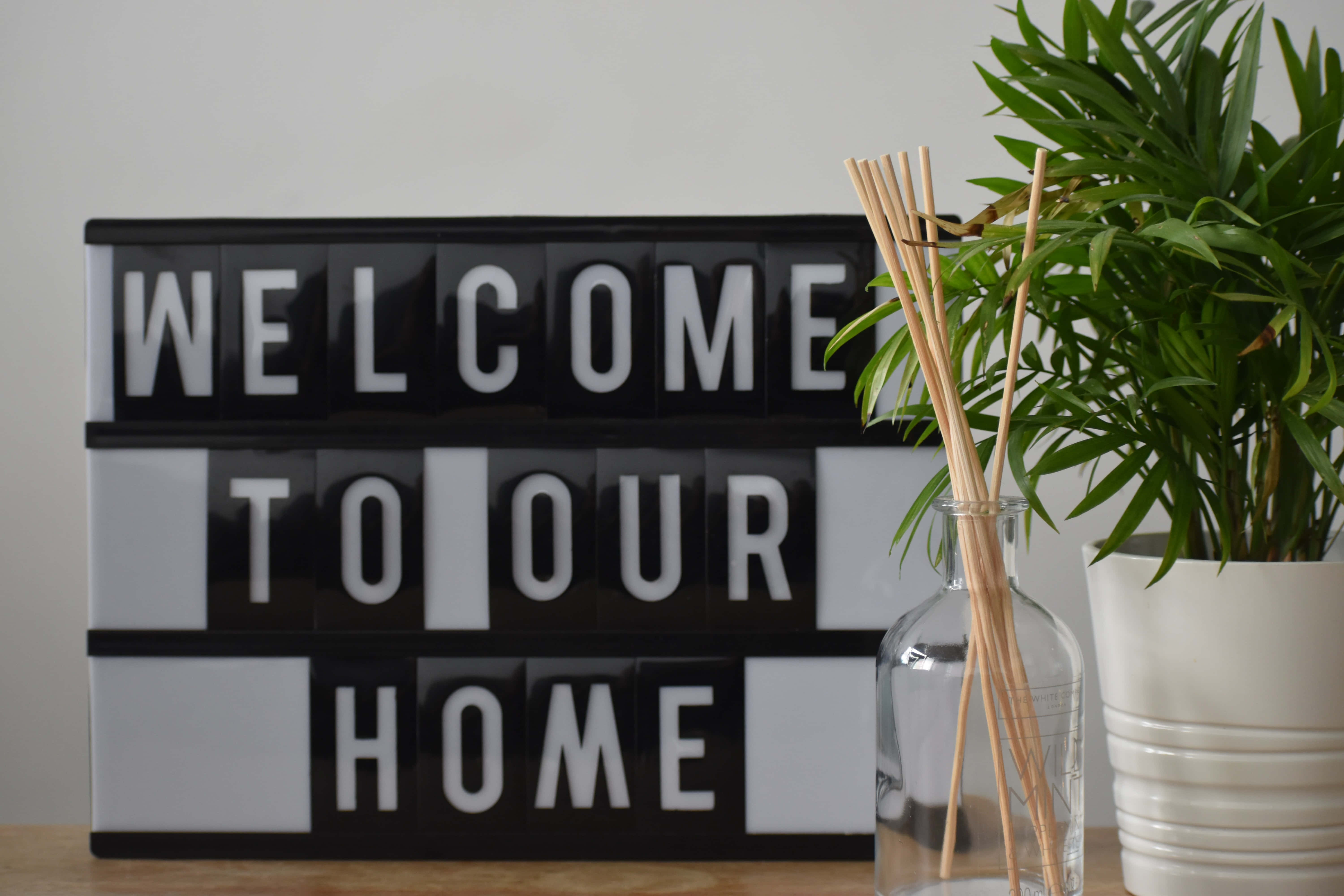 Welcome 桌布 壁紙 — 一個黑色燈箱，上面顯示「WELCOME TO OUR HOME」，旁邊放著盆栽植物和一個裝有蘆葦的玻璃花瓶，置於木質表面上。