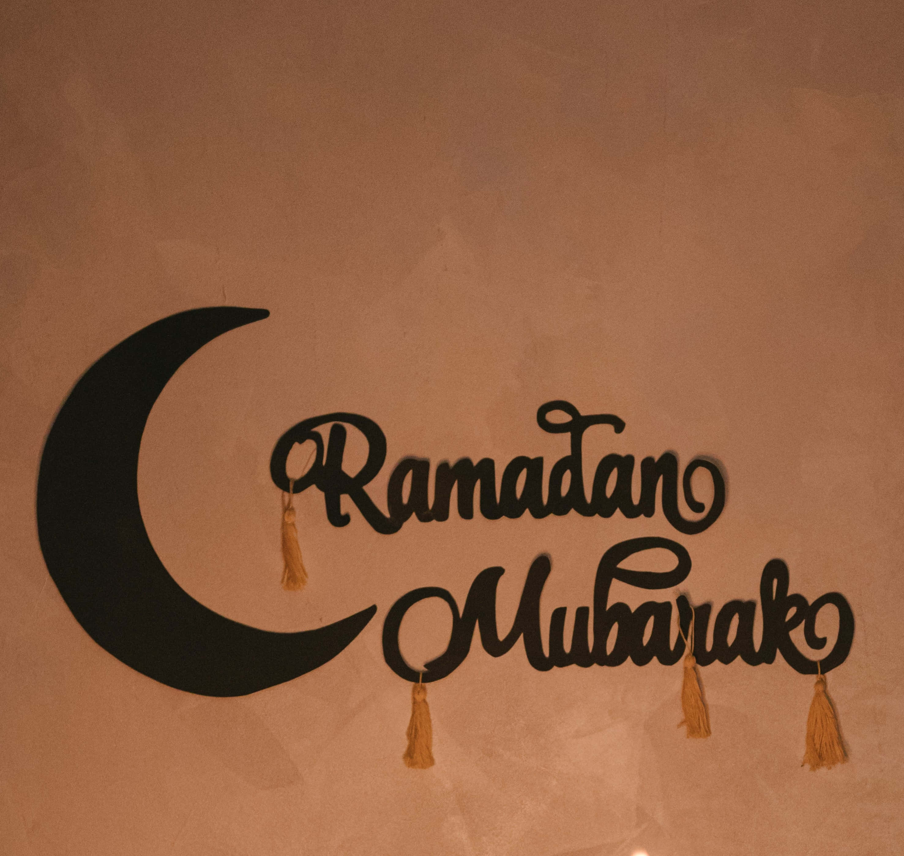 Ramadan Kareem 桌布 壁紙 — 一個黑色的新月和帶有流蘇的「Ramadan Mubarak」標誌掛在暖色調的紋理牆上。