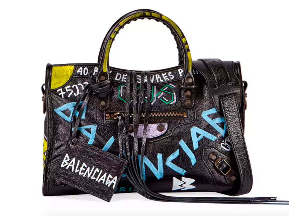 巴黎世家 桌布 壁紙 — 一個黑色皮革 Balenciaga 手提包，上面覆蓋著塗鴉風格的藍色、綠色和黃色文字及圖案，背景為白色。