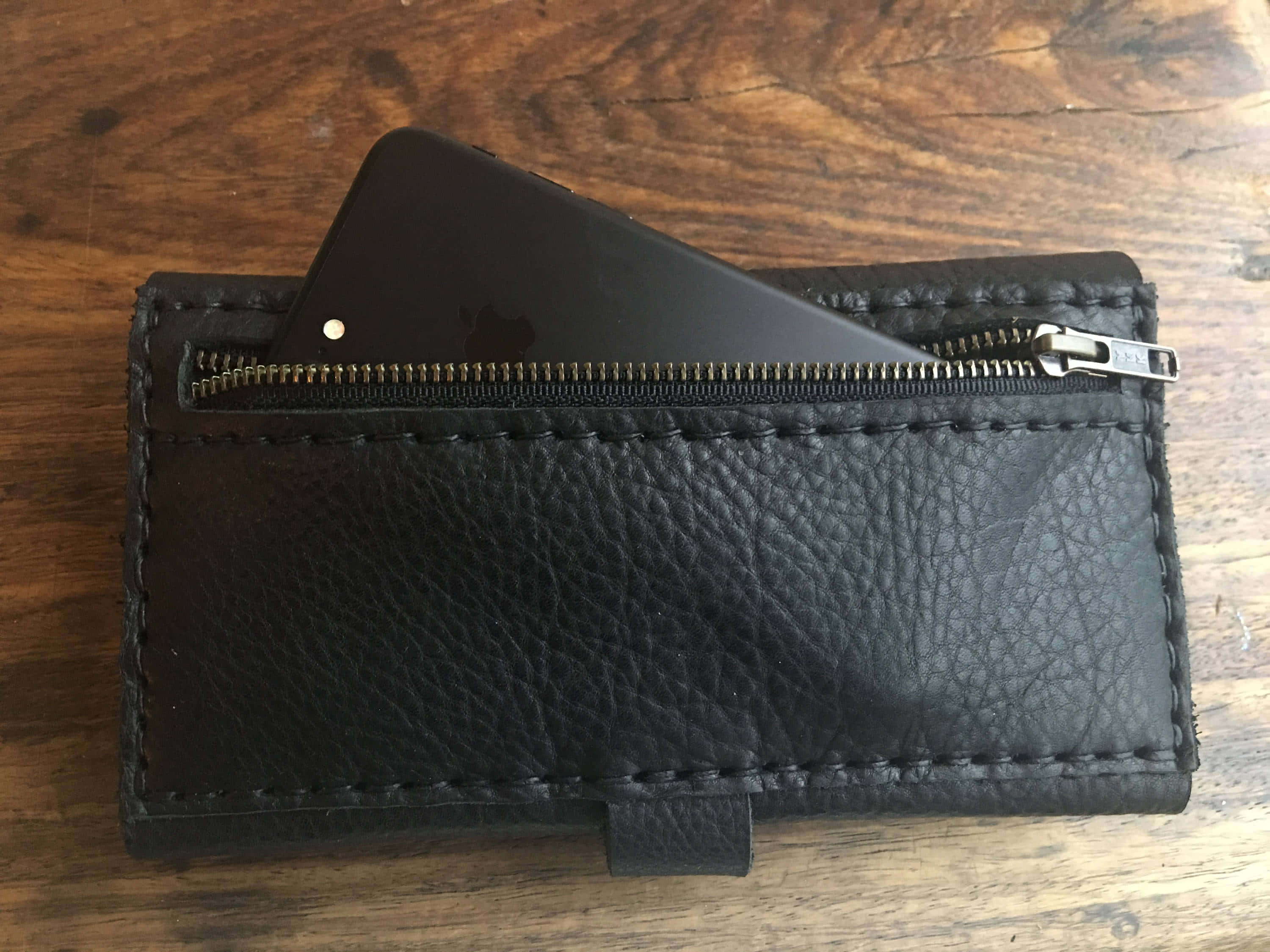 Black Leather 桌布 壁紙 — 一個黑色皮革錢包，拉鍊半開，露出一個黑色智慧型手機放在質樸的木質表面上。