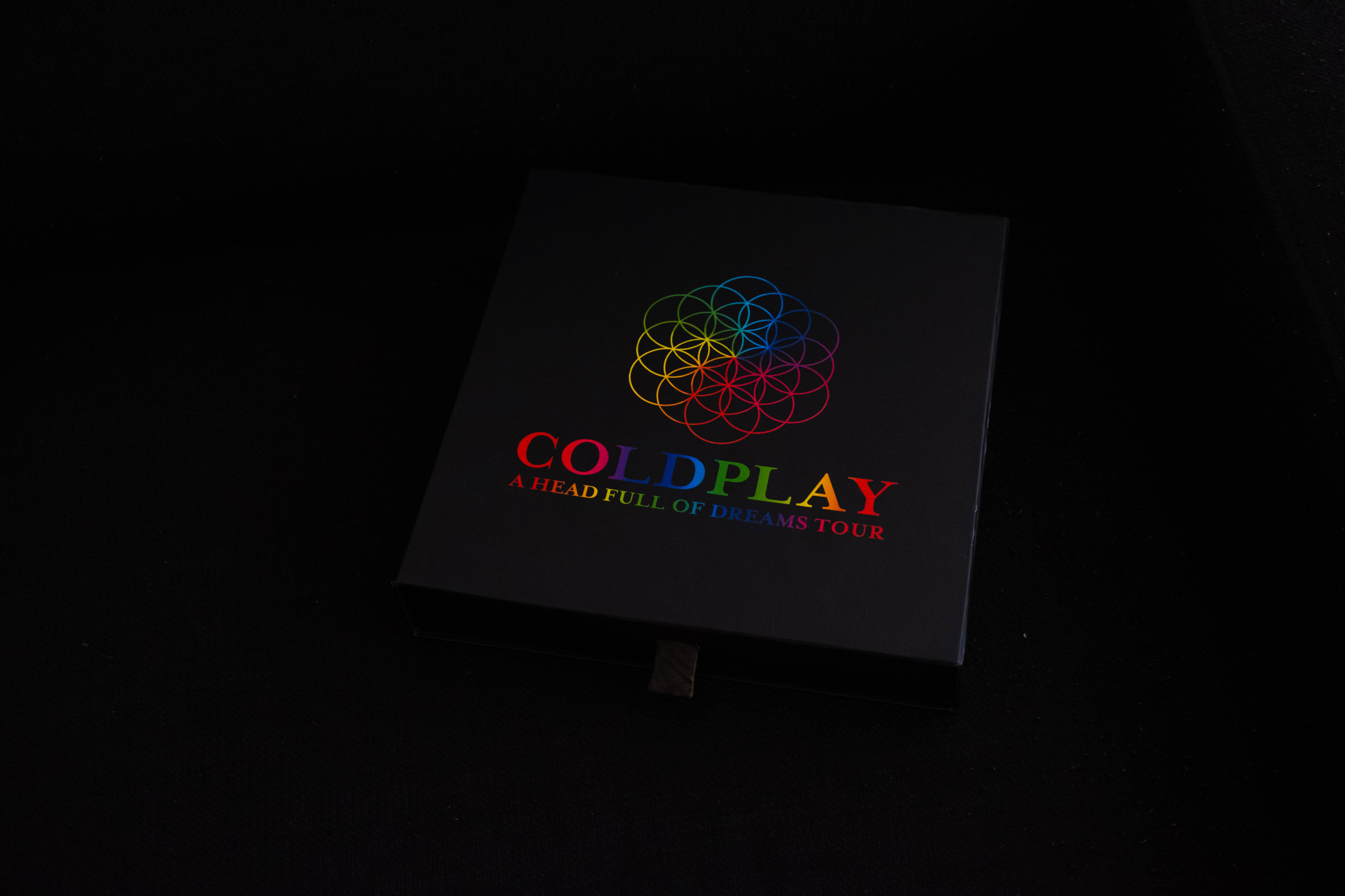 酷玩樂團 桌布 壁紙 — 一個黑色盒子，上面有Coldplay的「A Head Full of Dreams Tour」標誌和色彩繽紛的幾何花卉設計，居中放置在深色表面上。