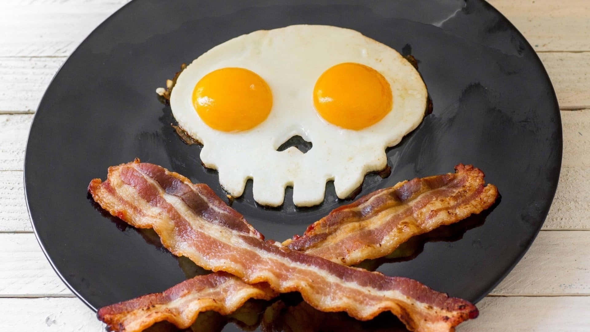 bacon 桌布 壁紙 — 一個黑色盤子上放著一個骷髏形狀的煎蛋，有兩個蛋黃作為眼睛，並以酥脆的培根條排列成交叉骨頭。
