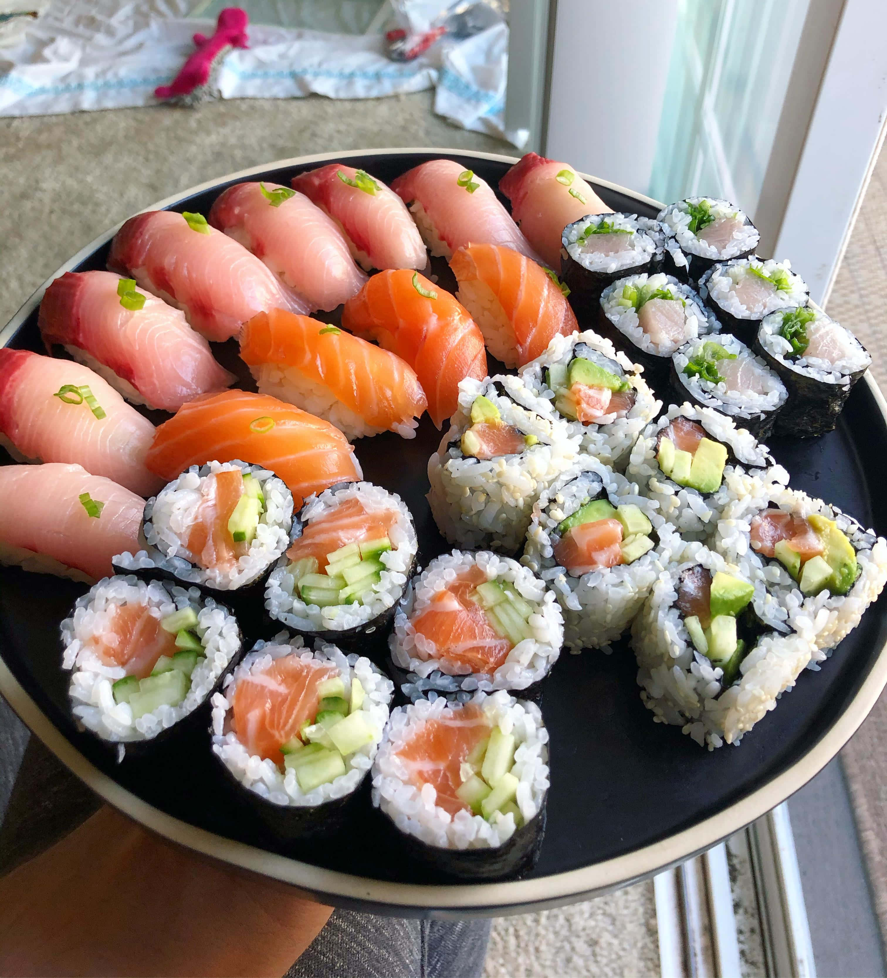 Sushi 桌布 壁紙 — 一個黑色盤子盛裝著令人垂涎的壽司拼盤，包括握壽司和卷壽司，上面放有鮭魚、鮪魚、酪梨和小黃瓜。