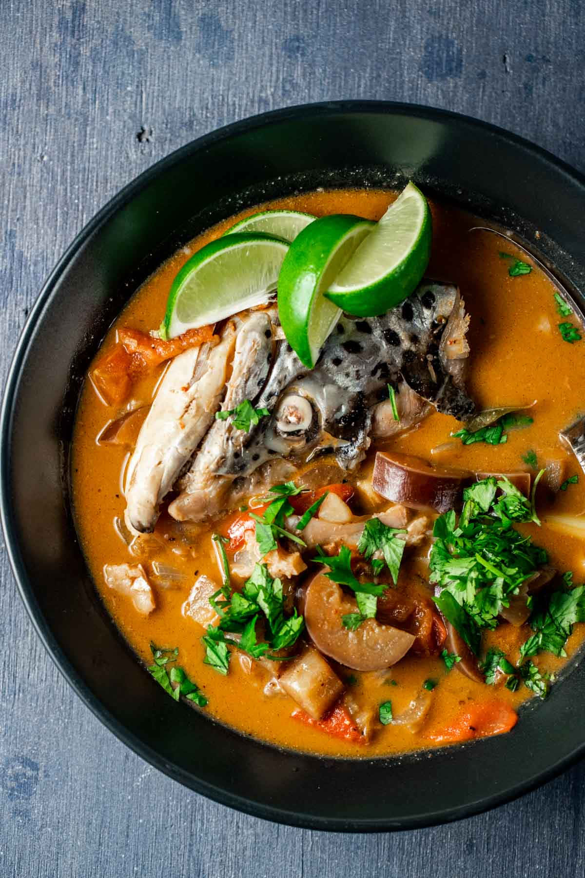 Food Photography 桌布 壁紙 — 一個黑色碗中的豐富橙色魚湯，上面放有萊姆角和新鮮香菜，置於有紋理的藍色表面上。
