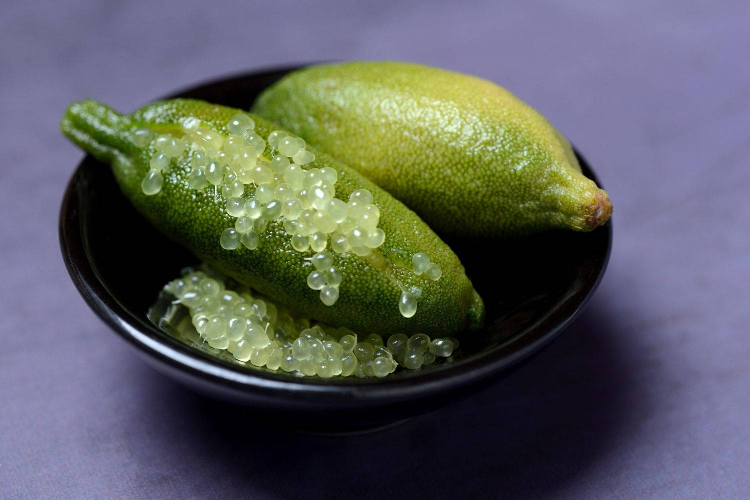 Fruits 桌布 壁紙 — 一個黑色碗裡有兩個充滿活力的綠色萊姆，其中一個上面綴有閃閃發光的半透明珍珠，背景為柔和的紫色。