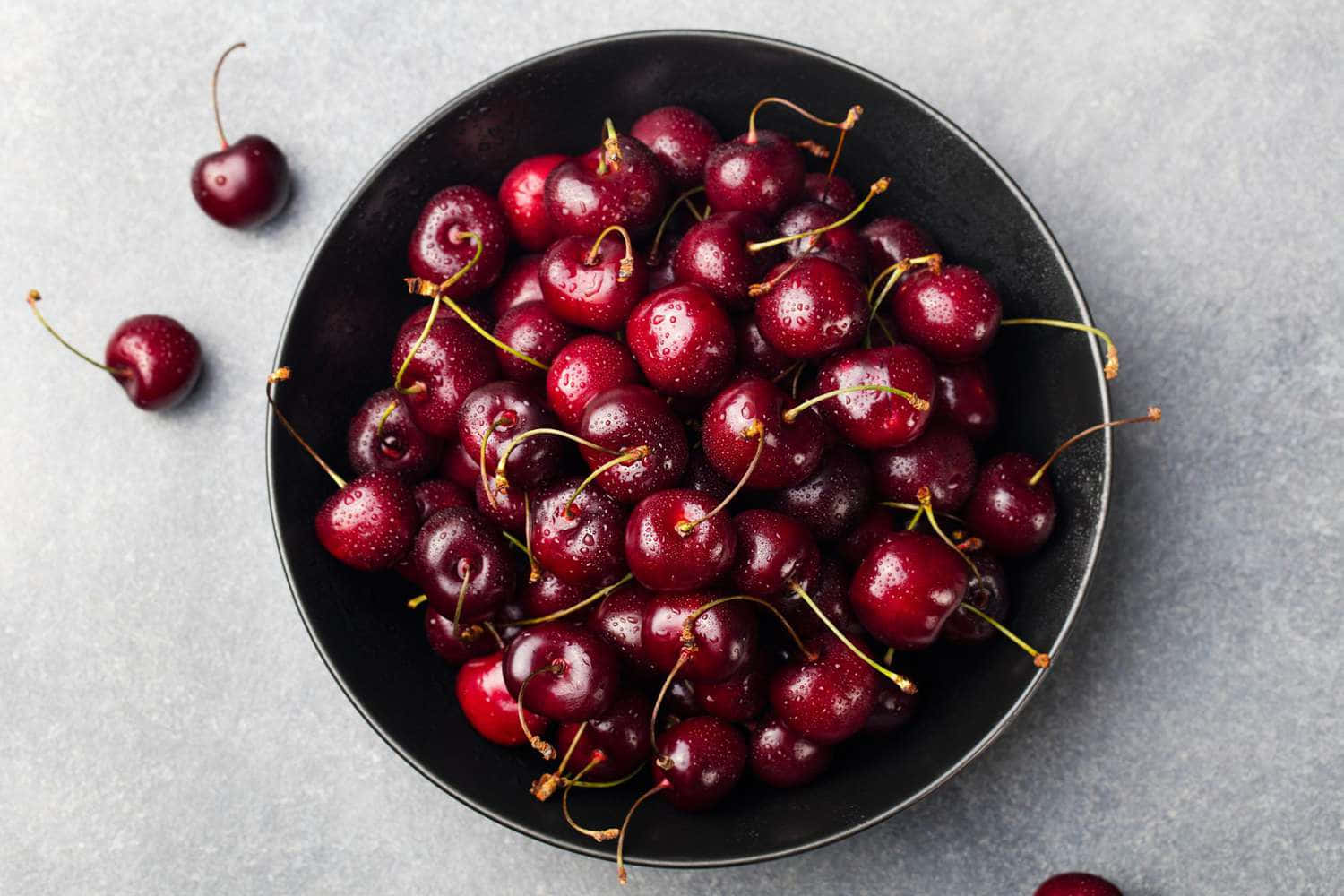 Red Cherries 桌布 壁紙 — 一個黑色碗裡裝滿了閃亮的紅色櫻桃，襯托著清涼的灰色石頭背景。