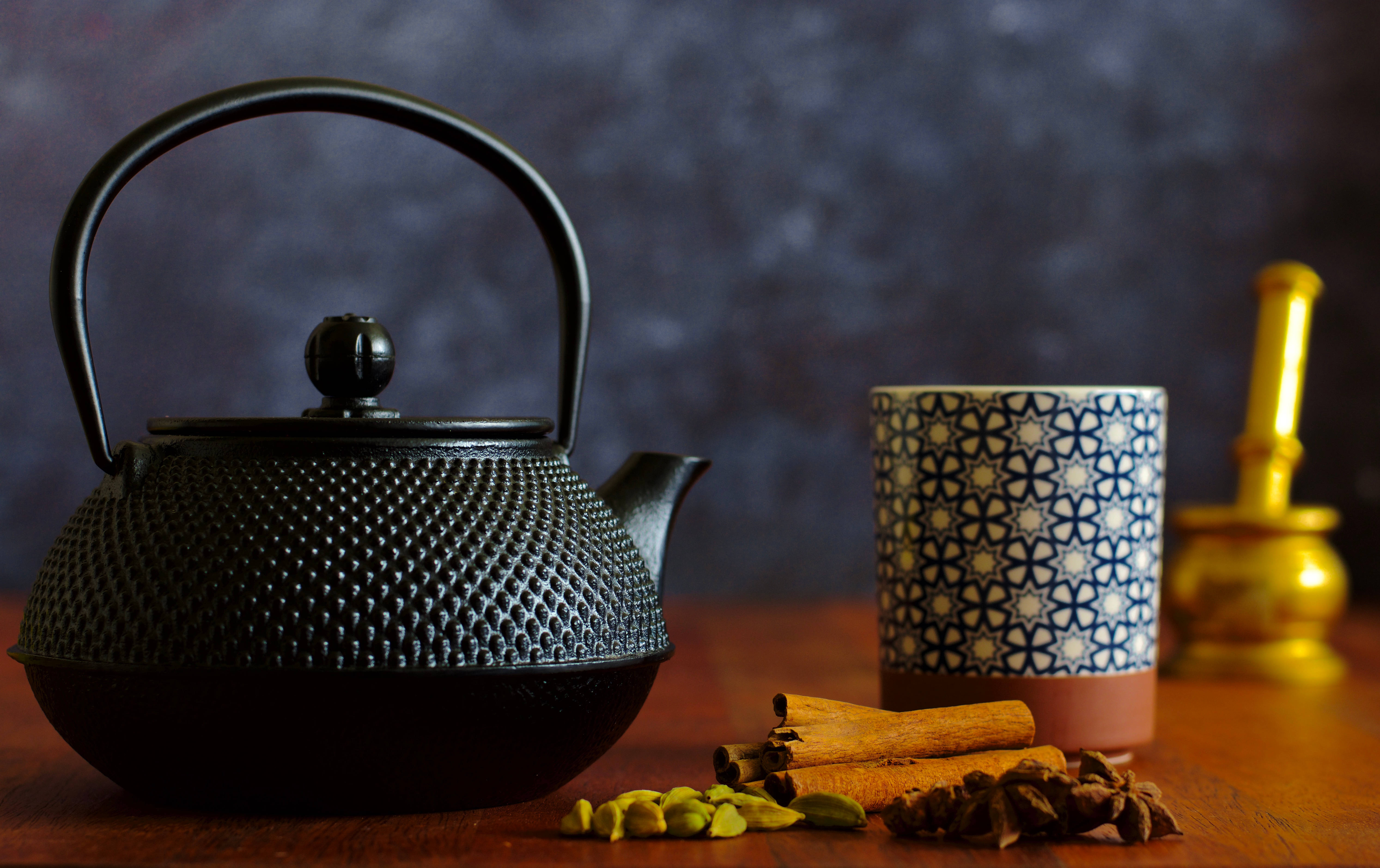 Rustic 桌布 壁紙 — 一個黑色紋理茶壺旁邊放著一個圖案杯子，杯中有肉桂棒和香料，放在木桌上，背景是深色紋理。