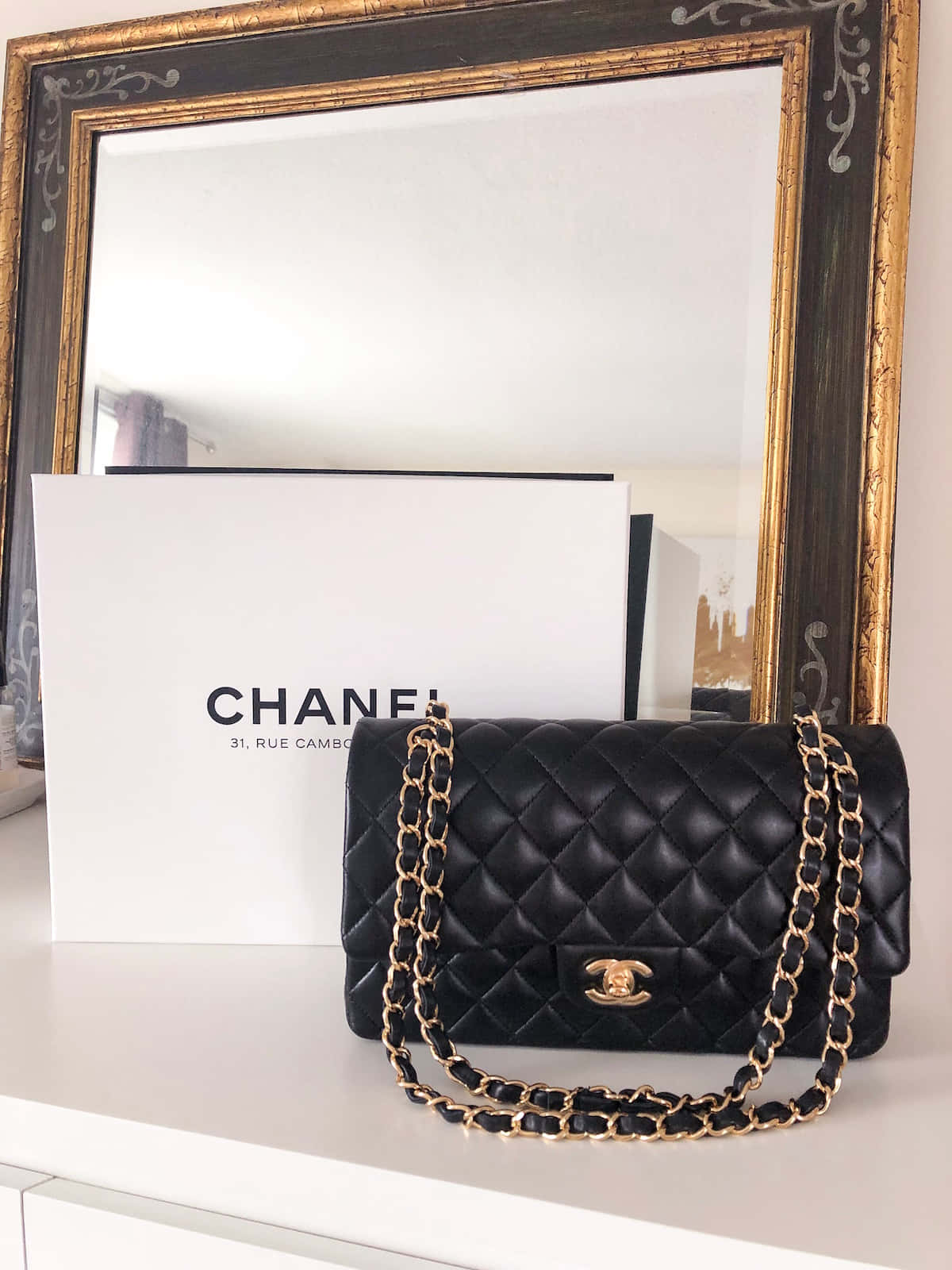 Chanel 桌布 壁紙 — 一個黑色絎縫Chanel手袋搭配金色鏈條，放置在白色表面上，旁邊是其品牌盒子和華麗鏡子。