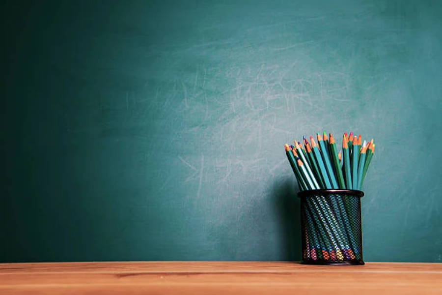 Education 桌布 壁紙 — 一個黑色網狀筆筒裝滿了色彩繽紛的鉛筆，放在一張木桌上，前面是一塊陳舊的綠色黑板。