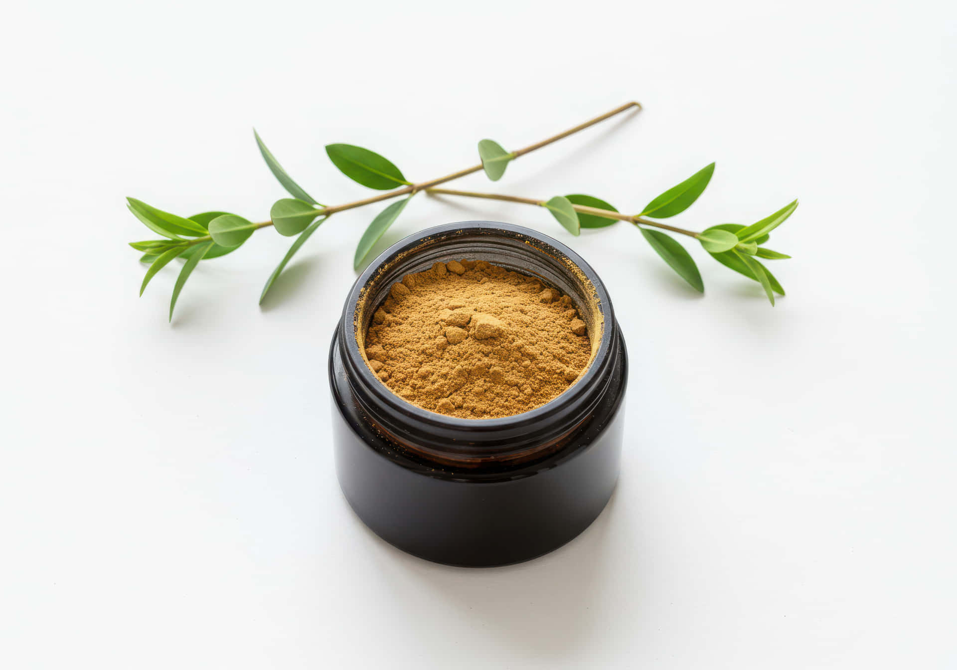 Natural cosmetic powder in a jar, isolated on white background 壁紙 — 一個黑色罐子裝滿金棕色粉末，旁邊放著一枝綠葉，在白色表面上。