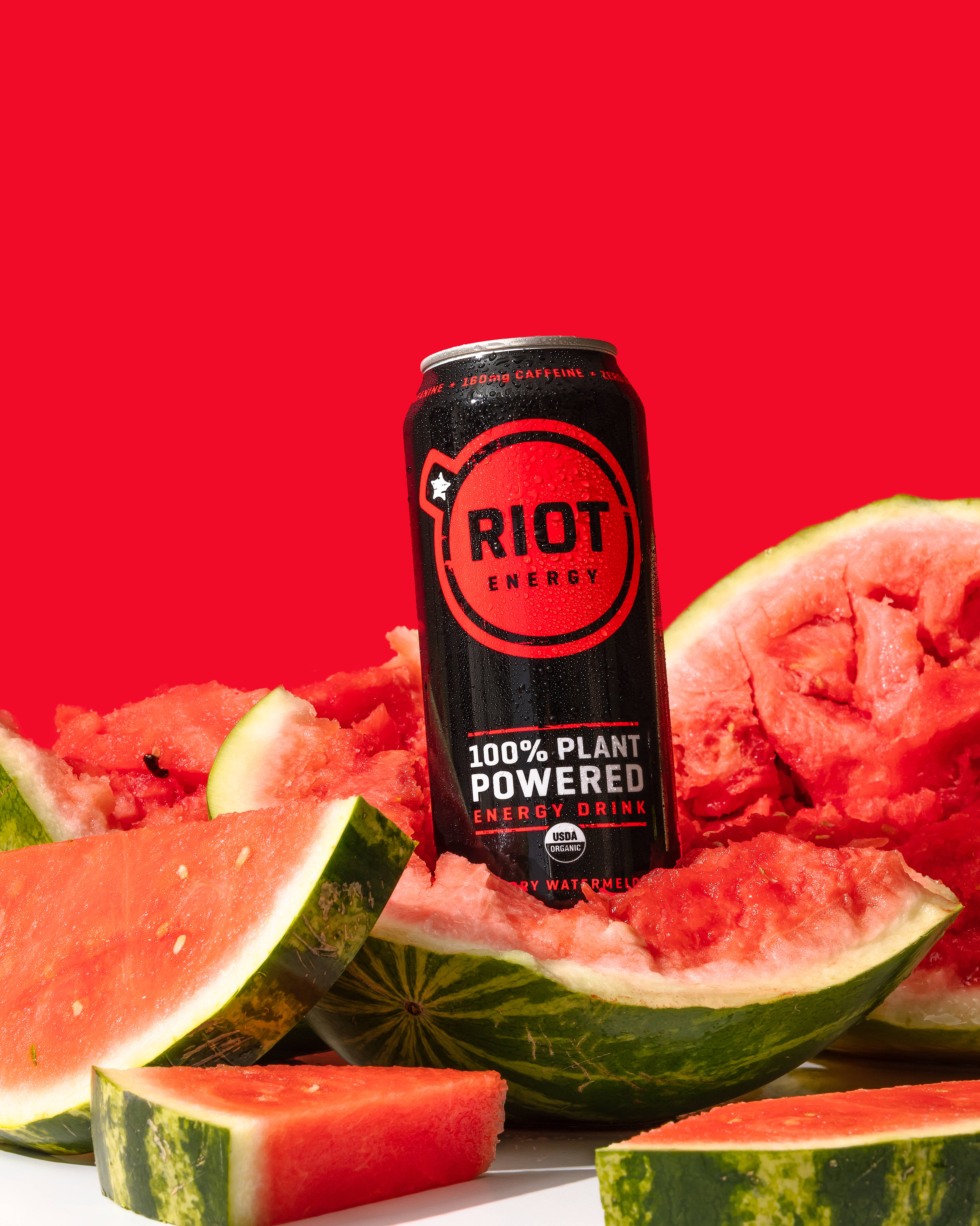 Watermelon 桌布 壁紙 — 一個黑色能量飲料罐，標有 'Riot Energy'，被多汁的西瓜片包圍，置於大膽的紅色背景上。