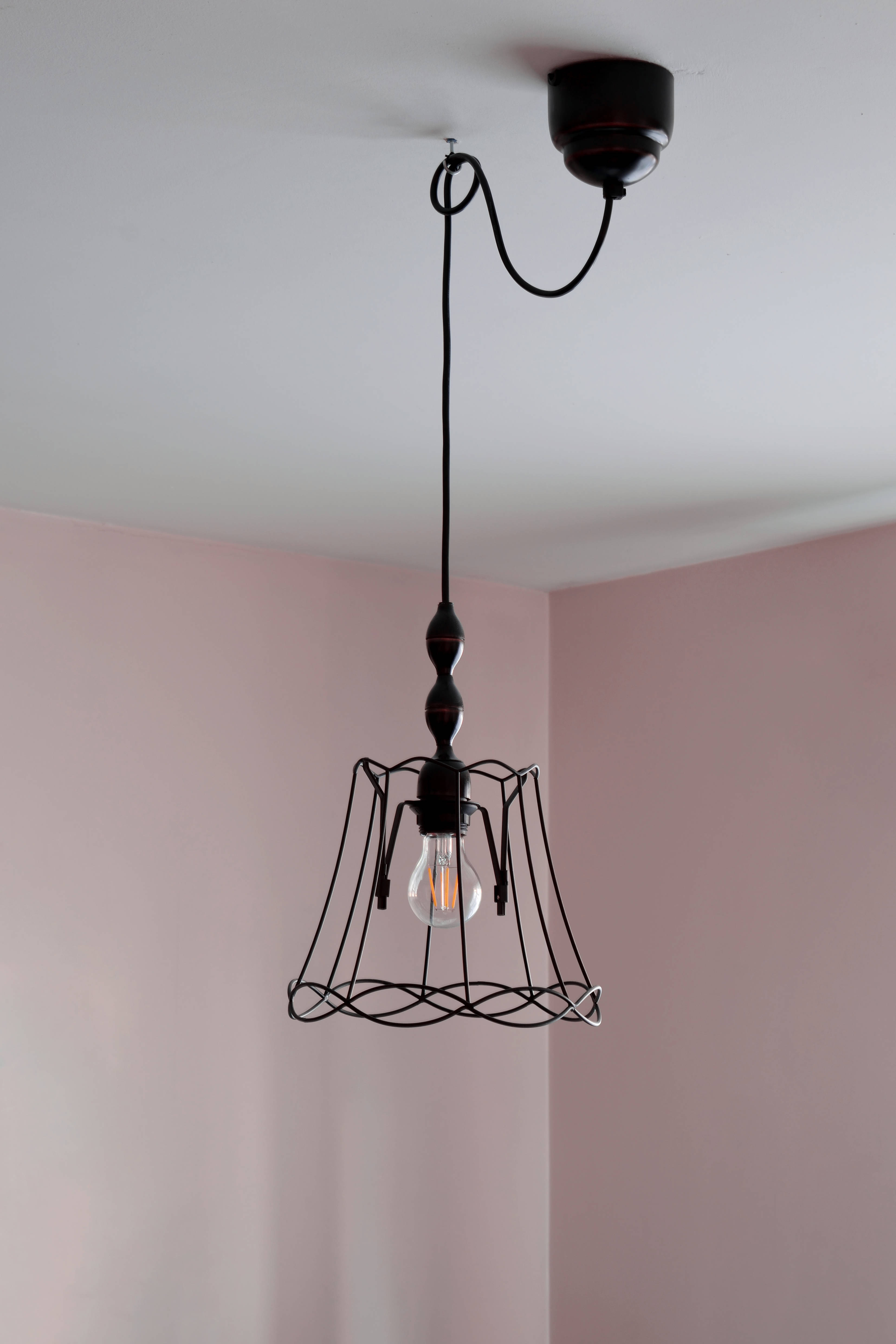 可愛女生 桌布 壁紙 — 一個黑色鐵絲籠吊燈，帶有裸露的燈泡，懸掛在彎曲的電線上，背景是柔和的粉紅色牆壁和白色天花板。