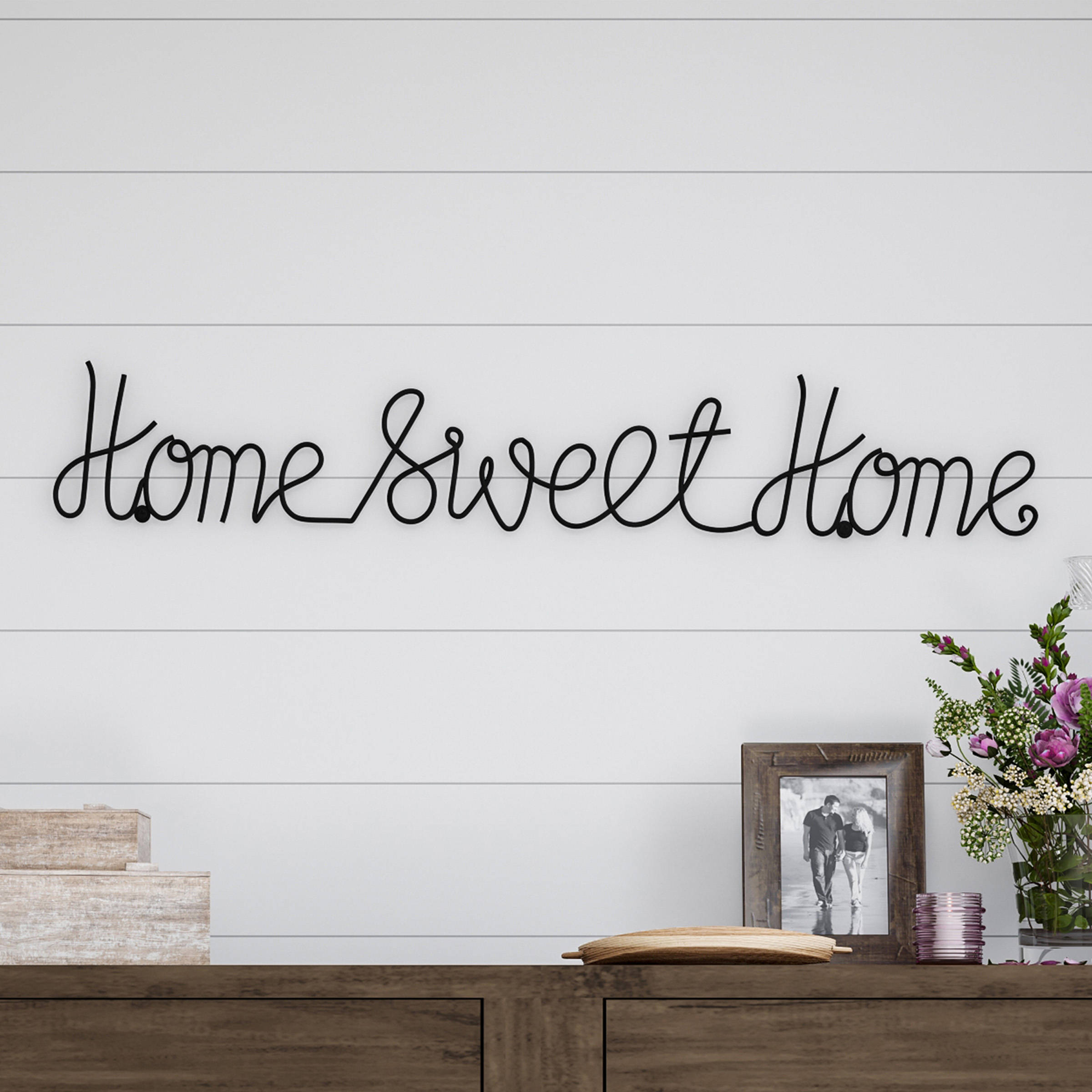 甜蜜的家 桌布 壁紙 — 一個黑色鐵線 'Home Sweet Home' 標牌掛在白色船艙牆上，下方木質矮櫃上放有相框和花卉擺設。