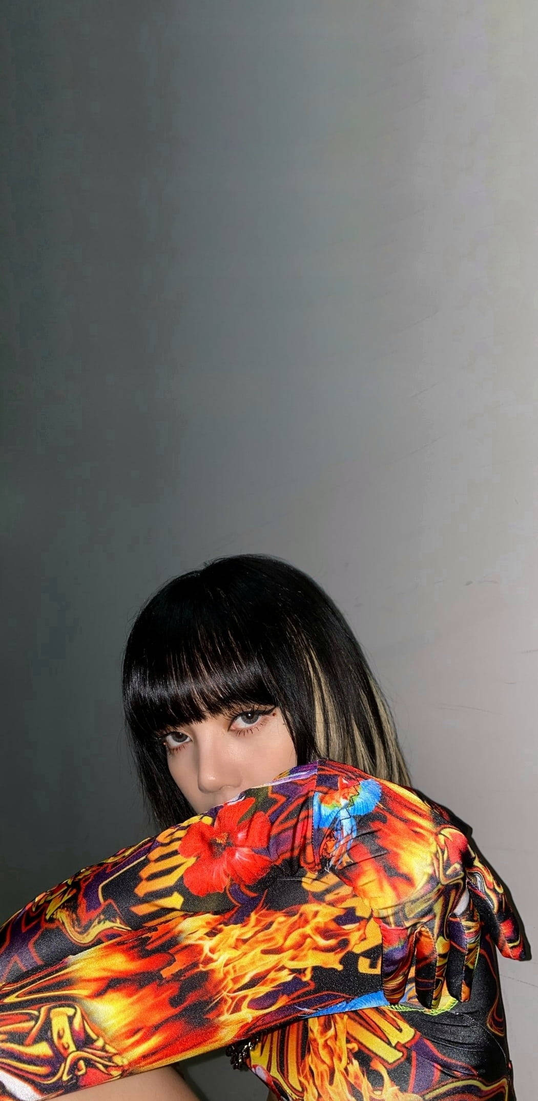 lisa 桌布 壁紙 — 一個黑色頭髮和瀏海的人從充滿活力的火焰圖案布料後面窺視，背景是純灰色牆壁。
