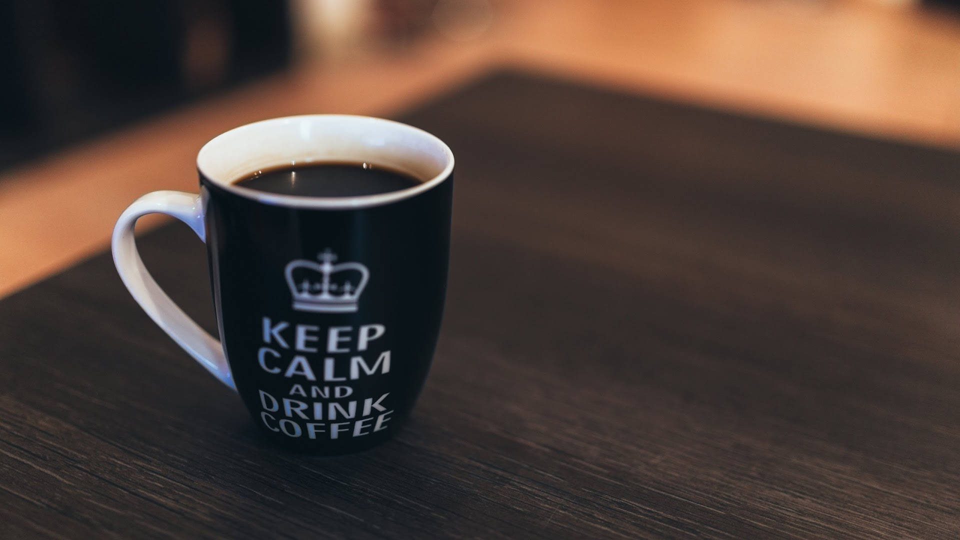 Coffee Aesthetic 桌布 壁紙 — 一個黑色馬克杯，上面有皇冠和「keep calm and drink coffee」字樣，放在深色木桌上，搭配溫暖的環境燈光。